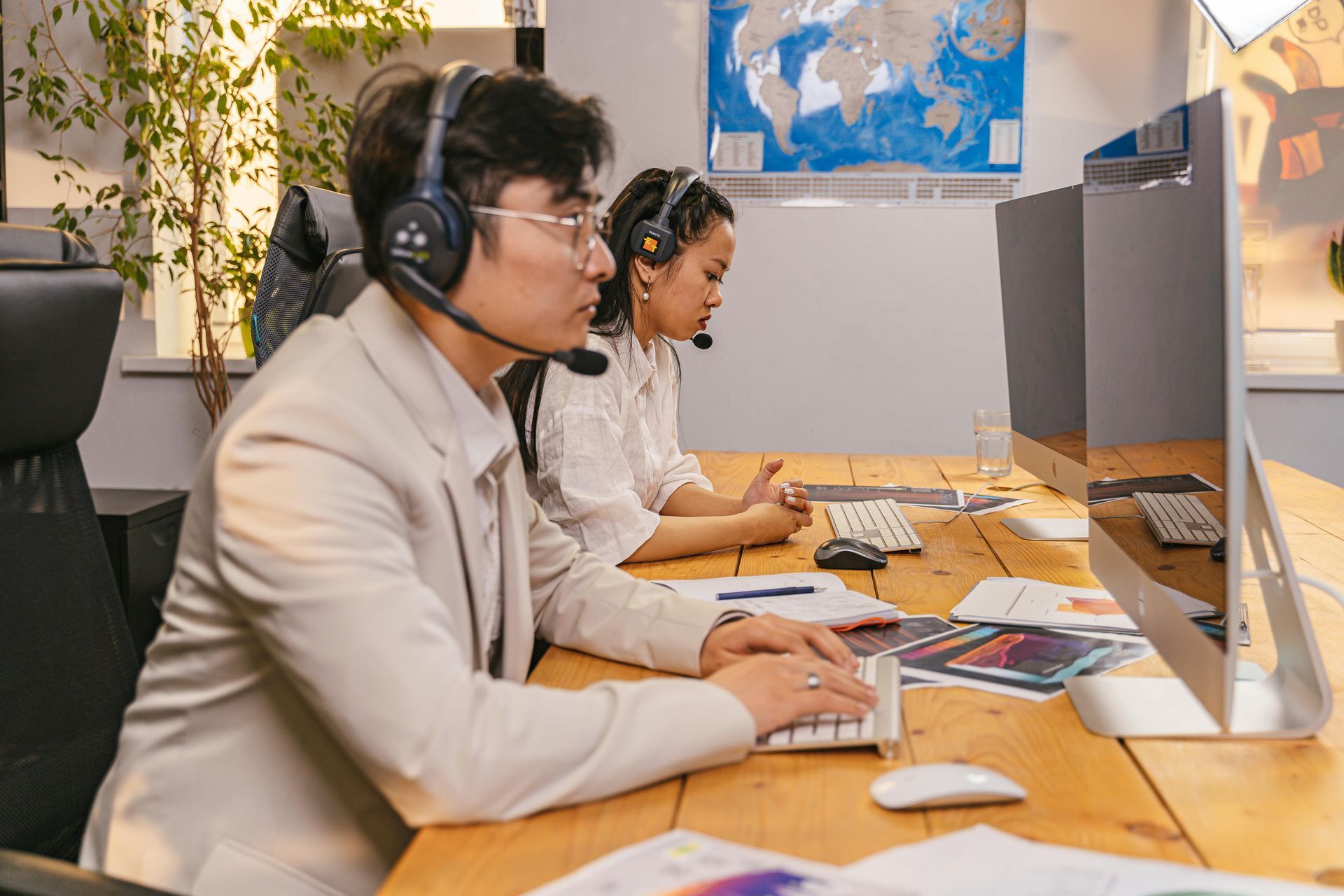 Two professionals wearing headsets work at computers in a bright office, seated at a long wooden desk with papers.