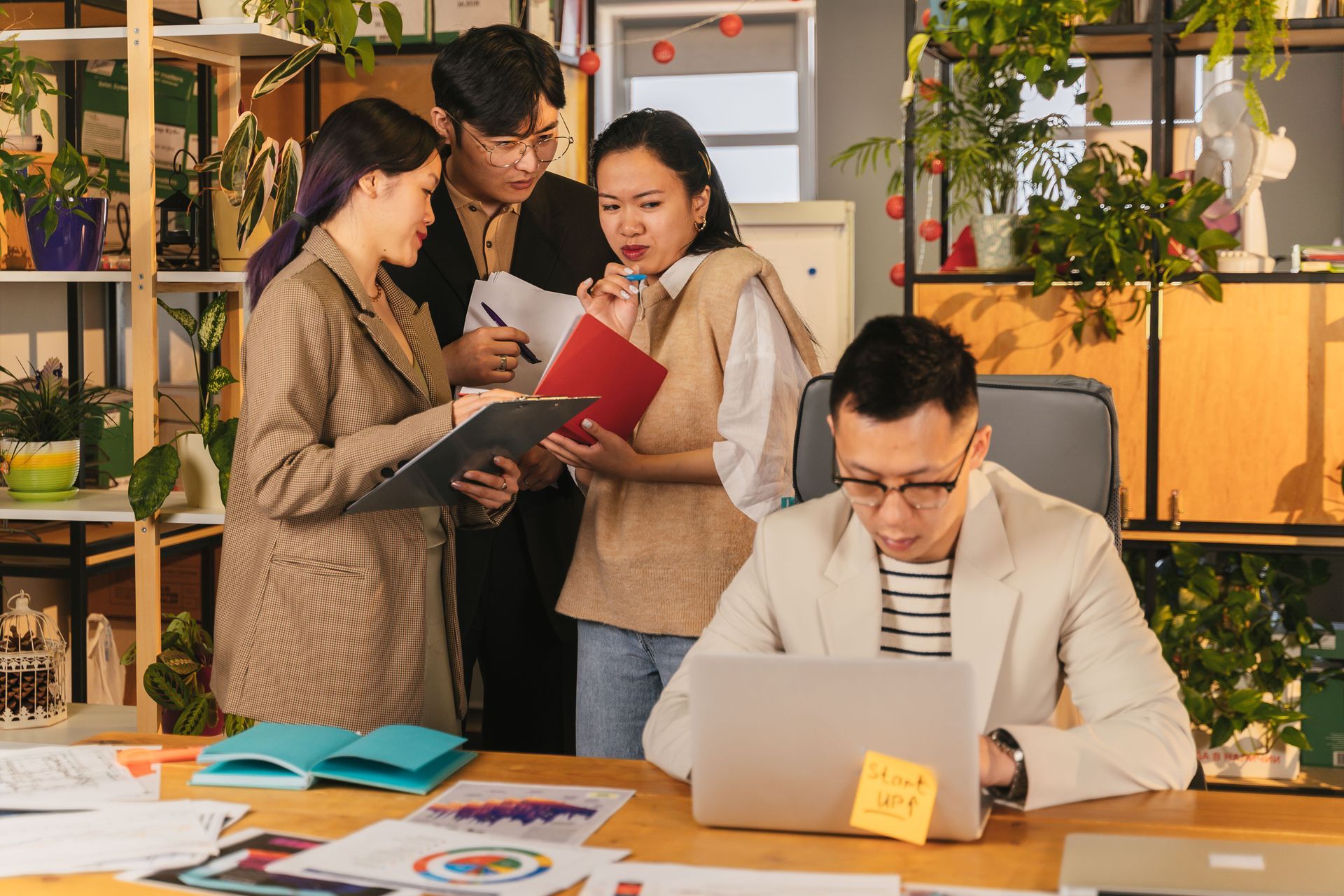 Four coworkers discuss documents while one works at a laptop in a bright office.