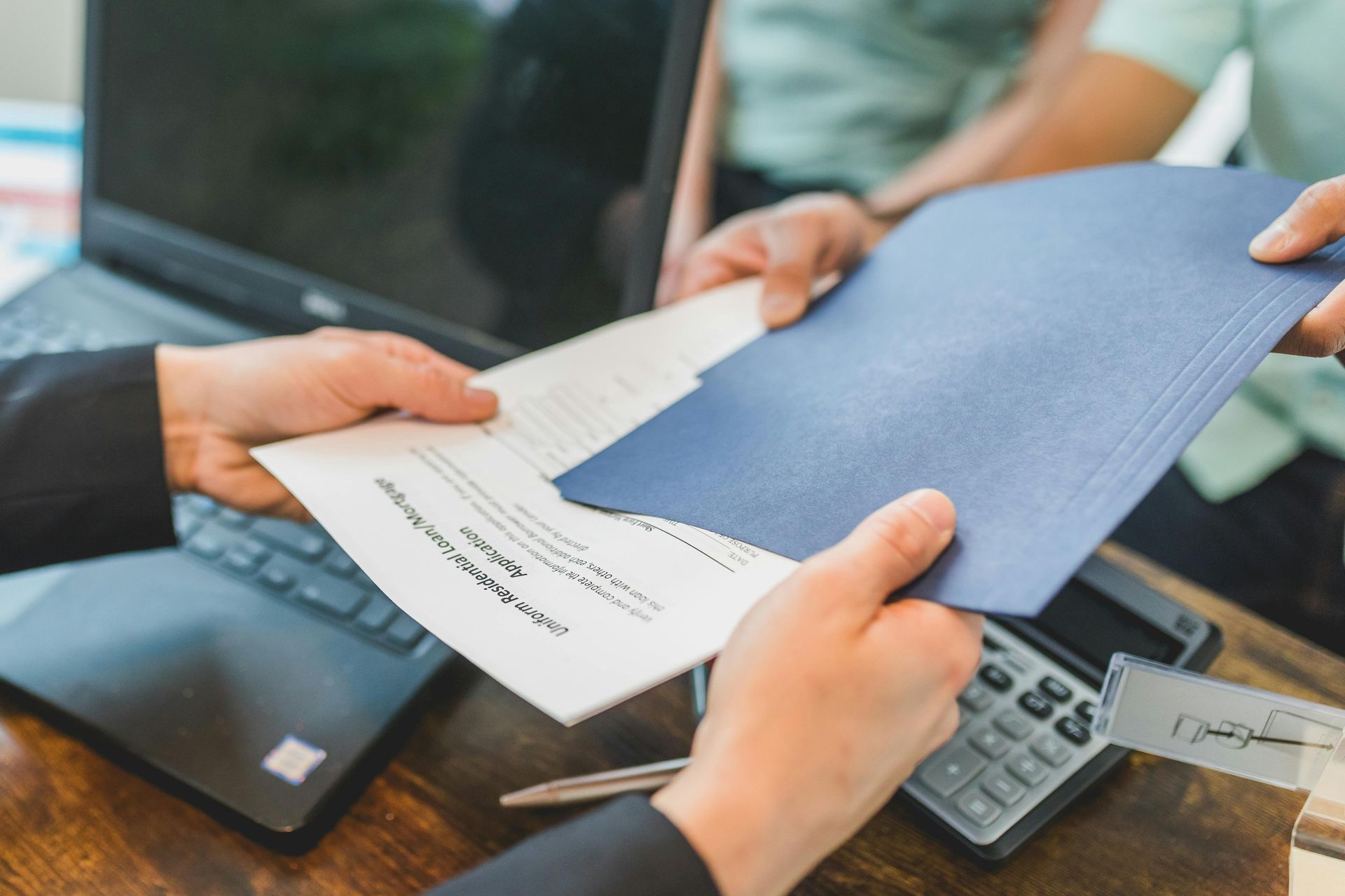 Hands exchange documents over a wooden desk with a laptop and calculator, indicating a professional meeting.