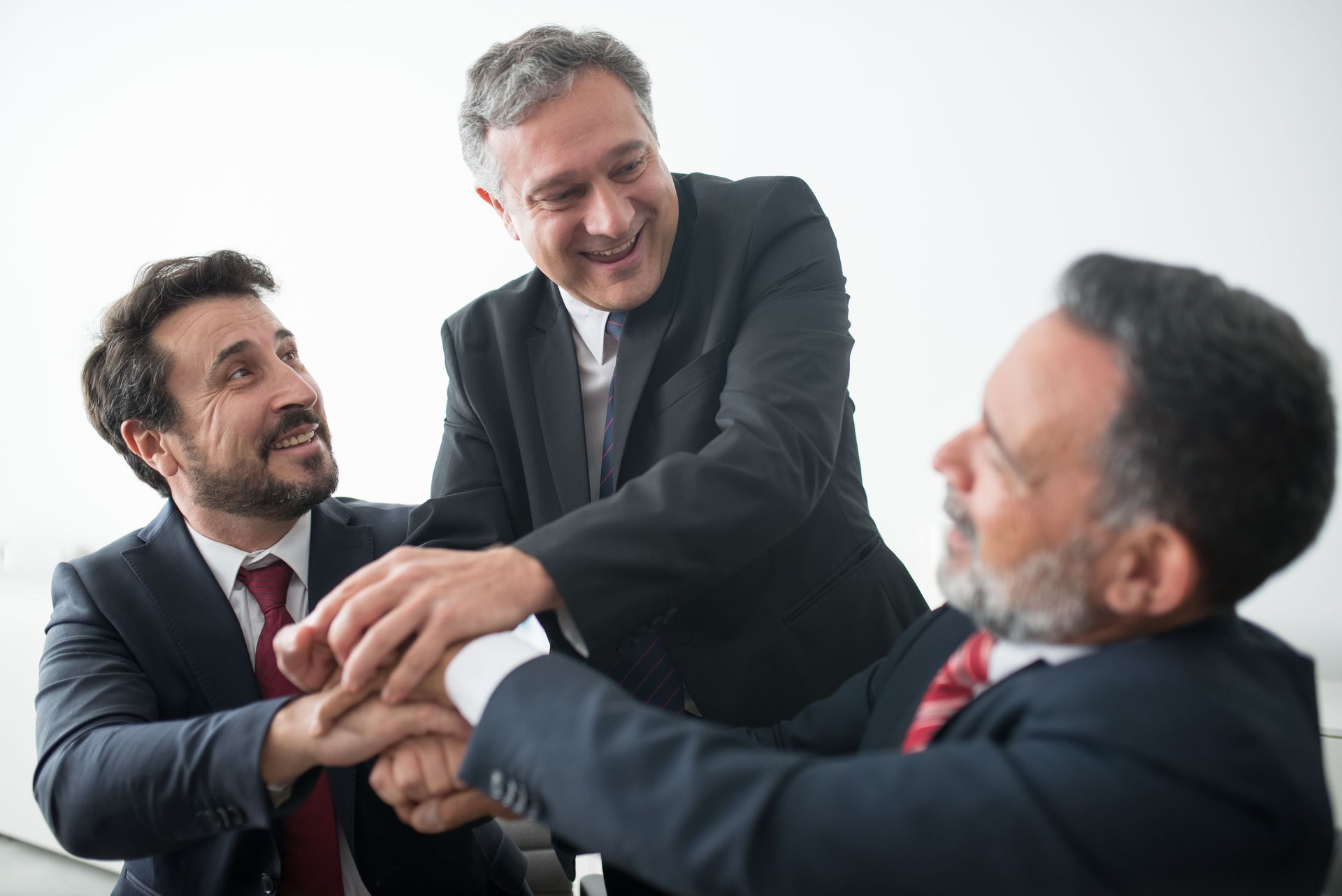 Three business people in suits stack hands in a meeting, smiling in a bright office.