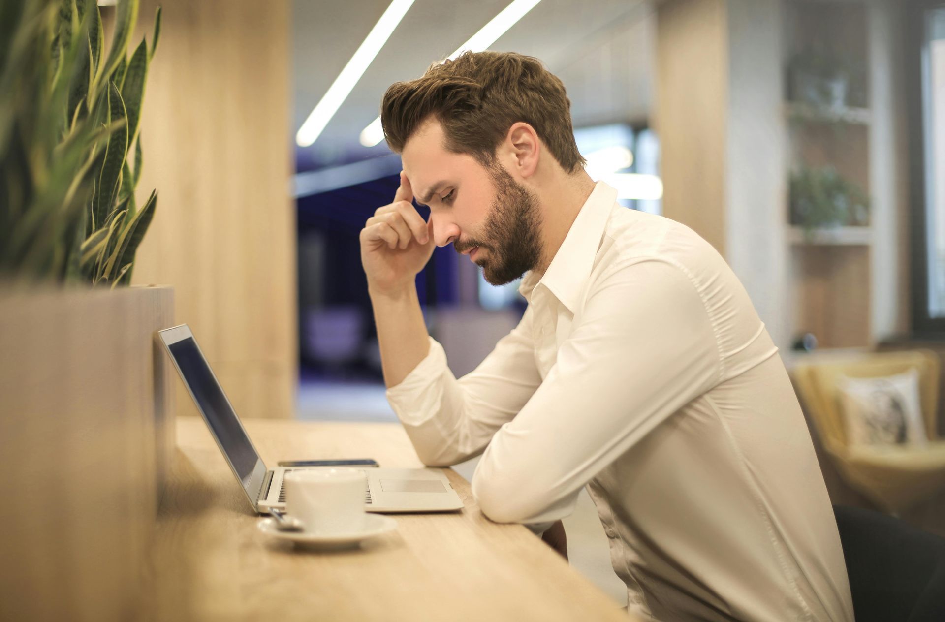 A person in a white button-down shirt sits at a desk with a laptop, looking stressed while resting their head on their hand.