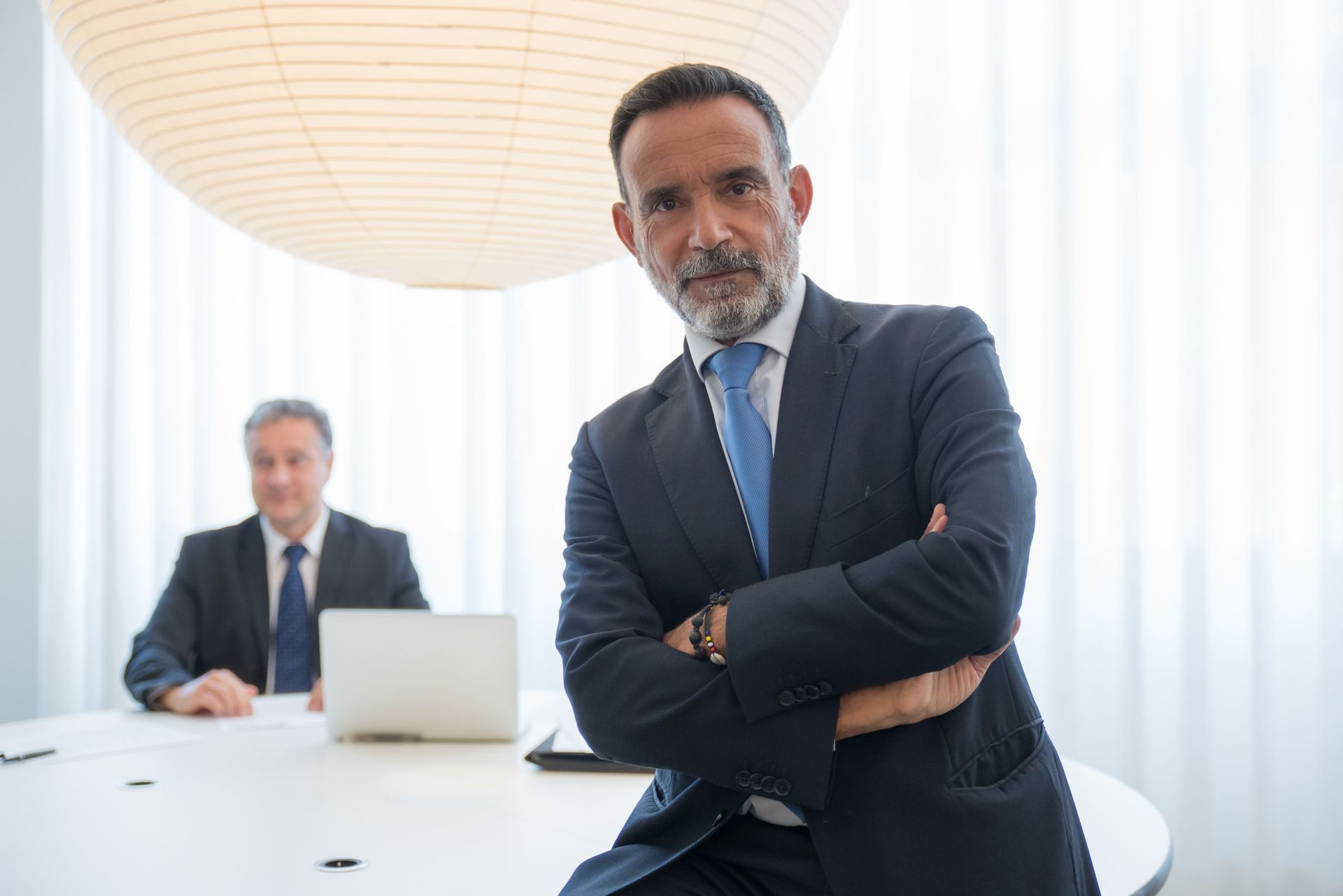 Business professionals in a bright conference room, one standing with arms crossed, one seated with a laptop.