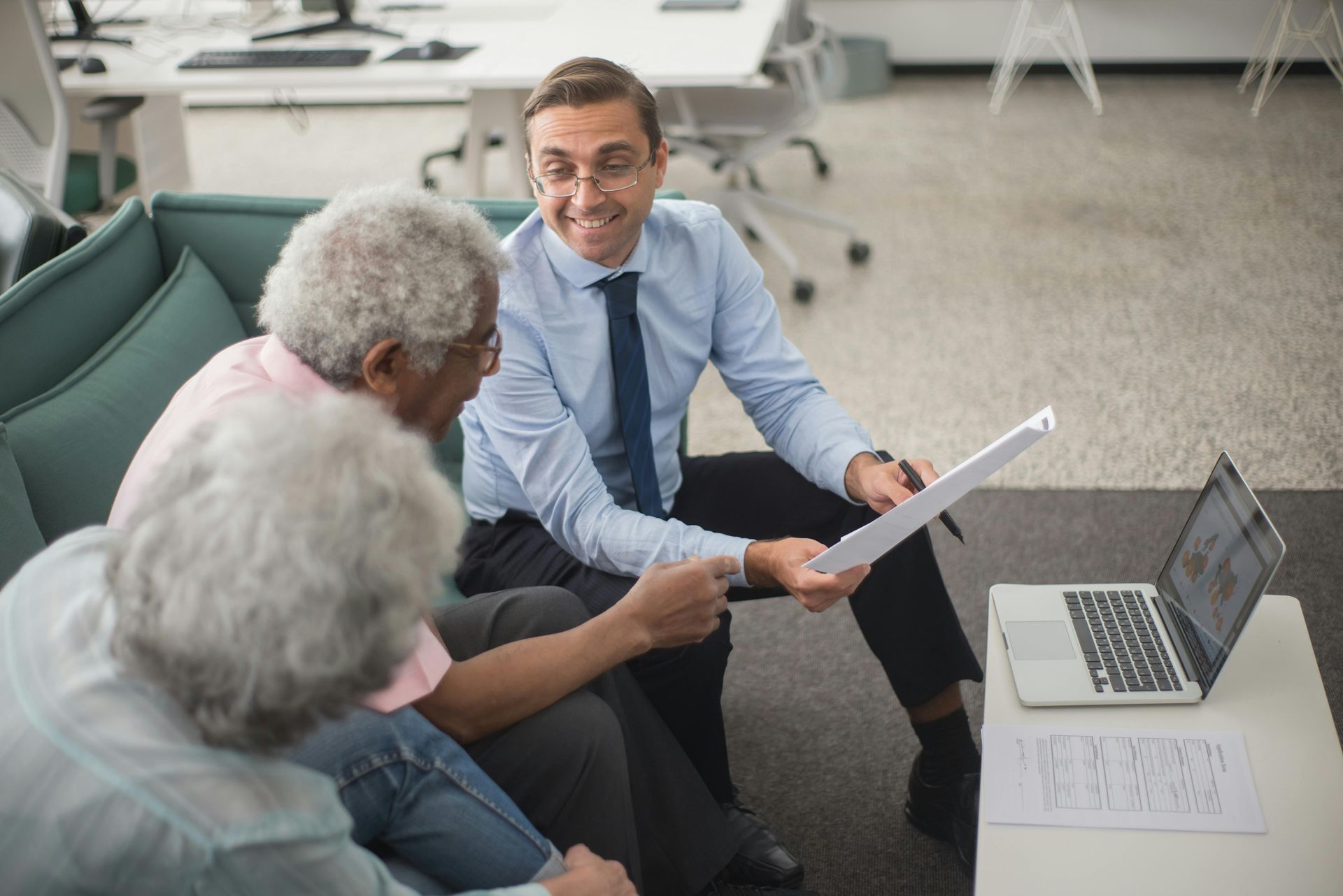 Financial advisor showing documents to two older adults seated on a sofa. Laptop and papers on table.