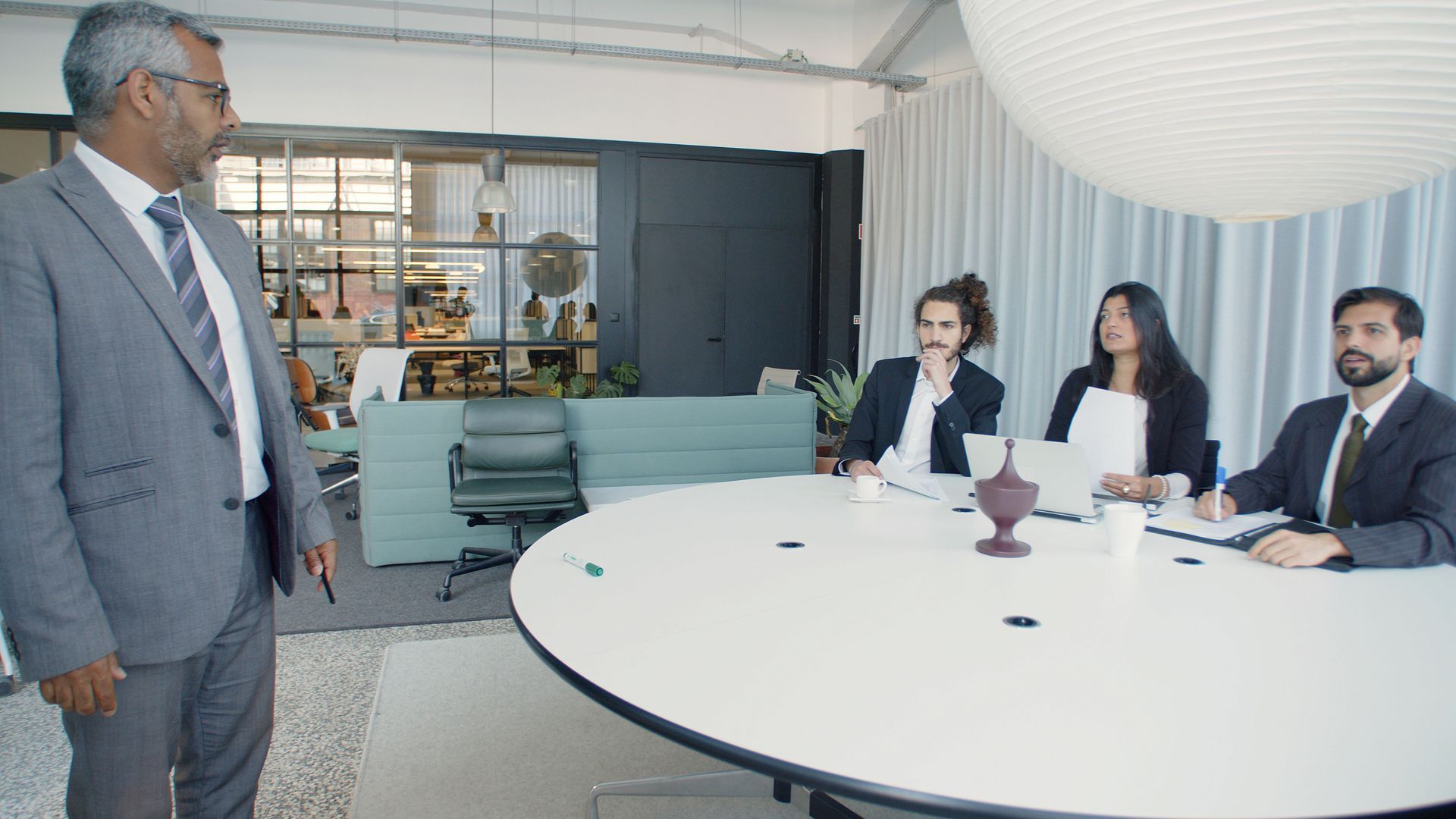 Business meeting in a bright conference room, with one man speaking to three seated colleagues around a round table.