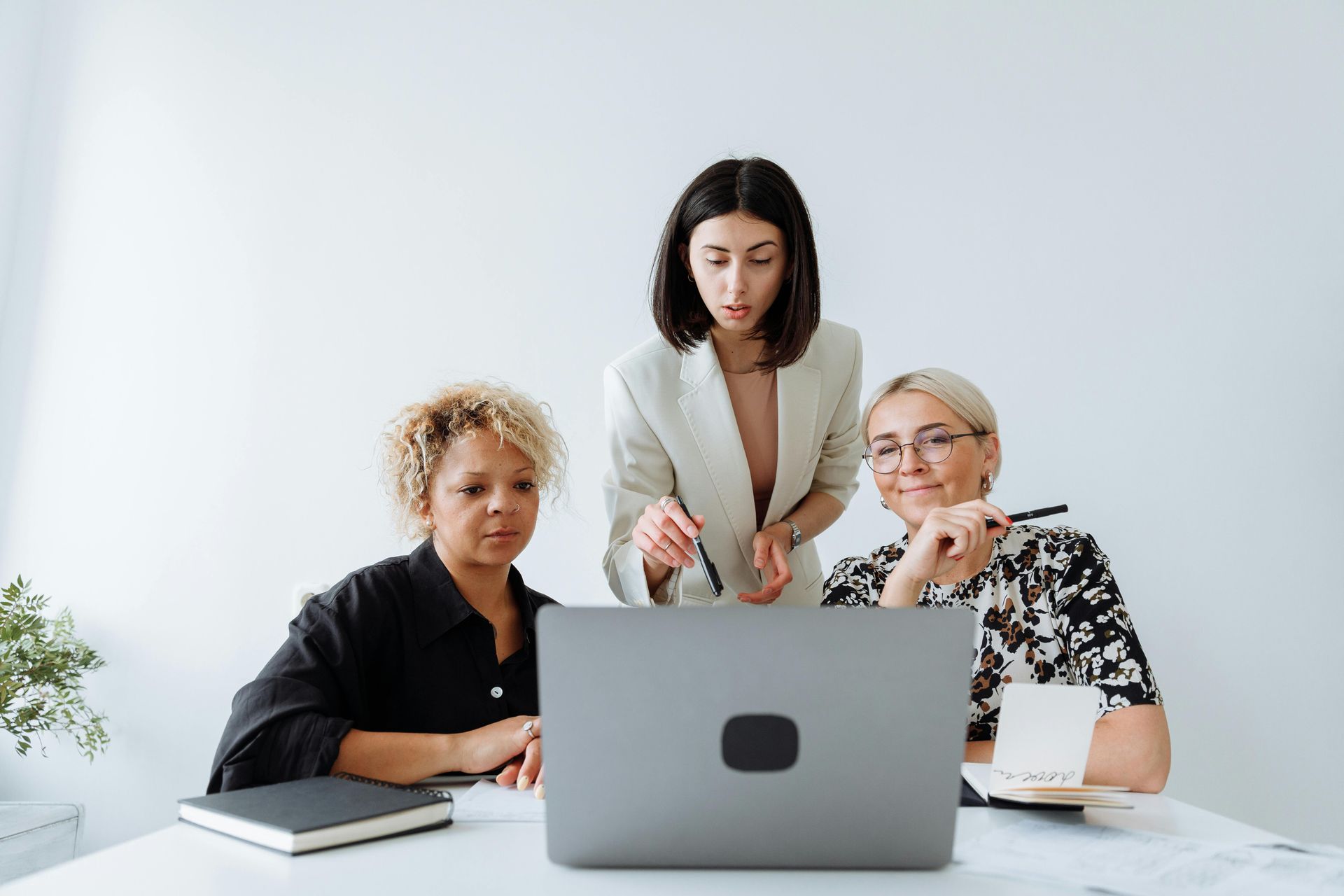 Three people collaborating around a laptop on a white desk in a minimalist, brightly lit office space.