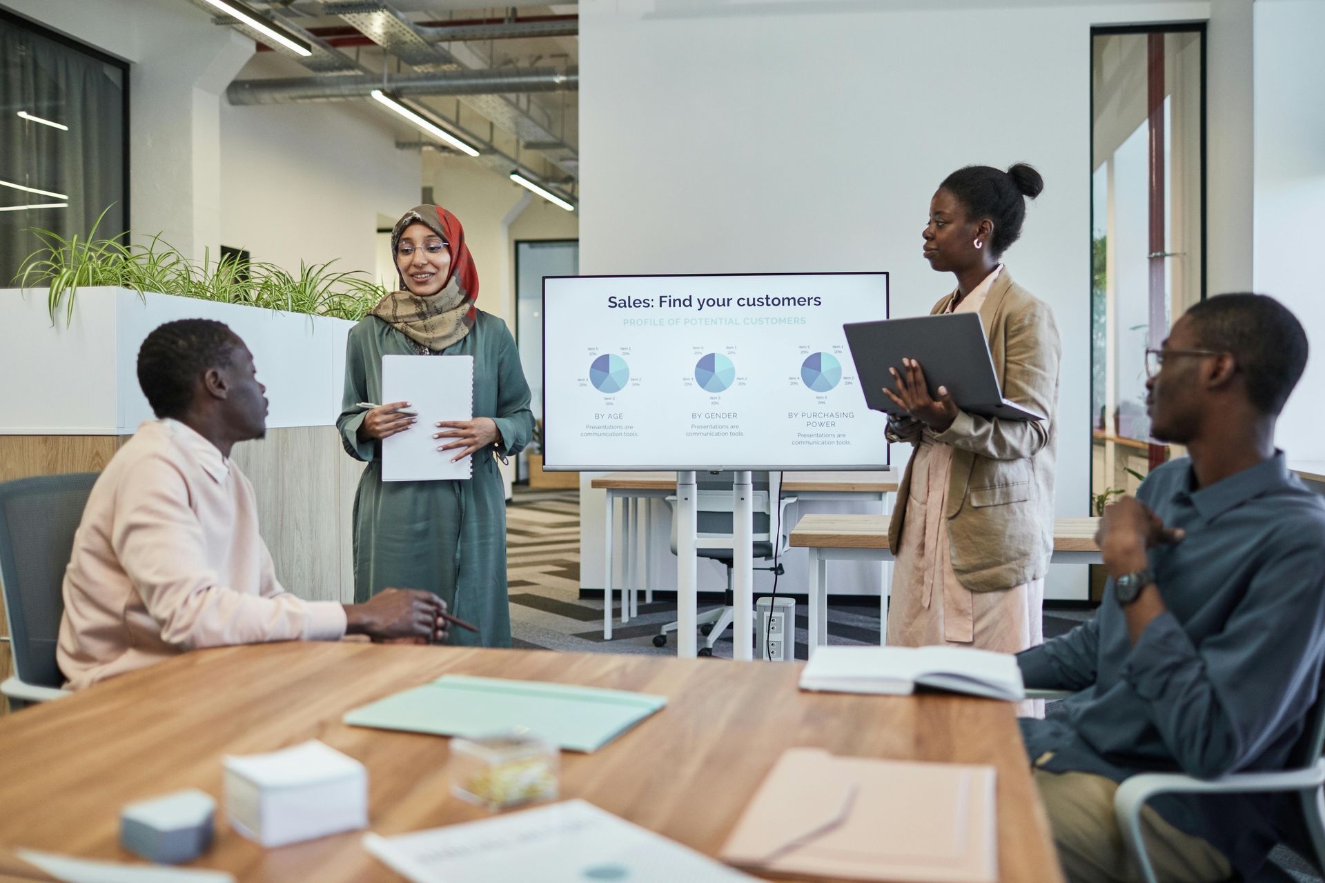 Four professionals collaborate in an office, with two standing by a screen showing business charts while others listen.