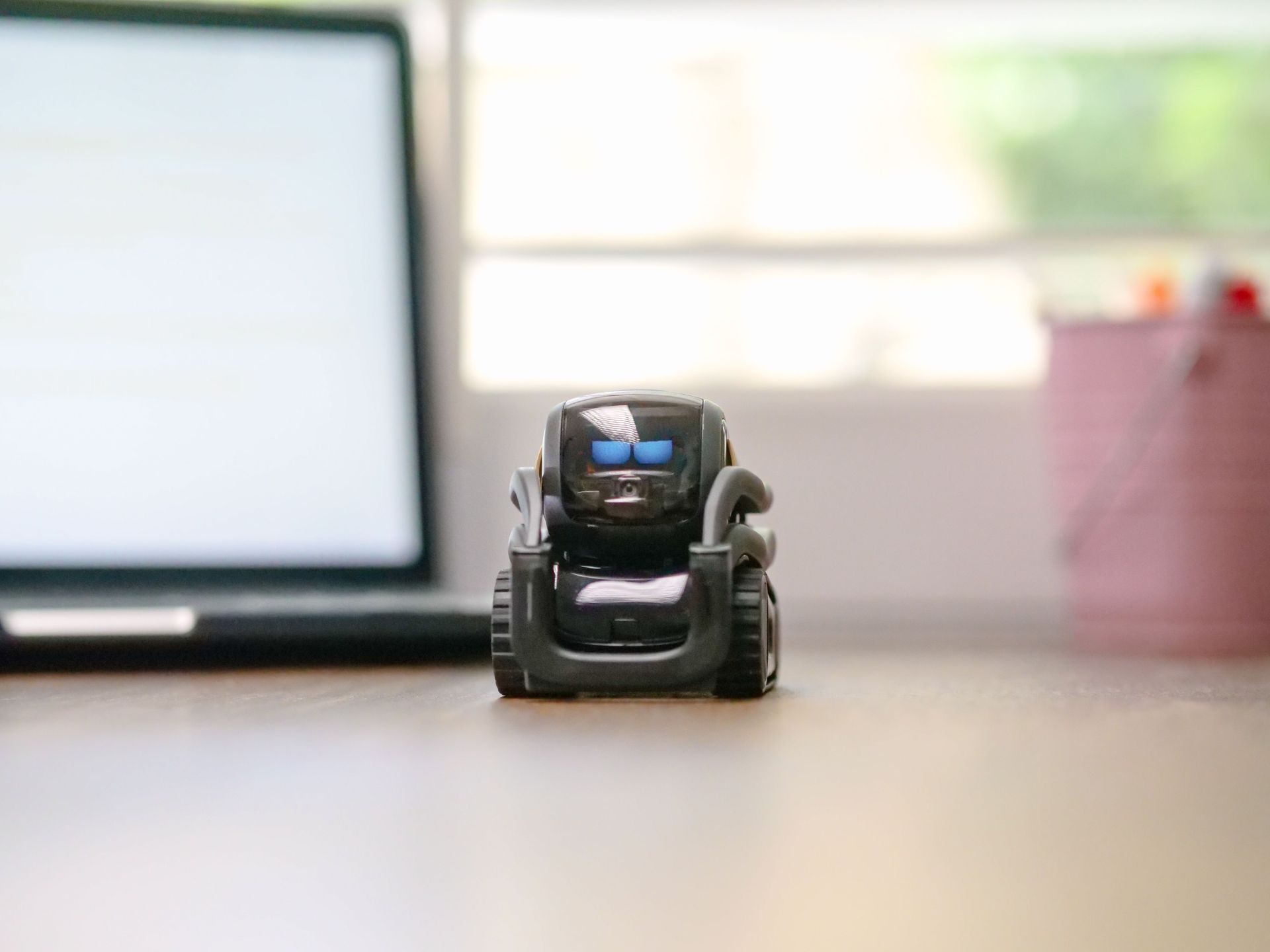 Small toy robot on a desk with a blurred laptop and pink cup in the background
