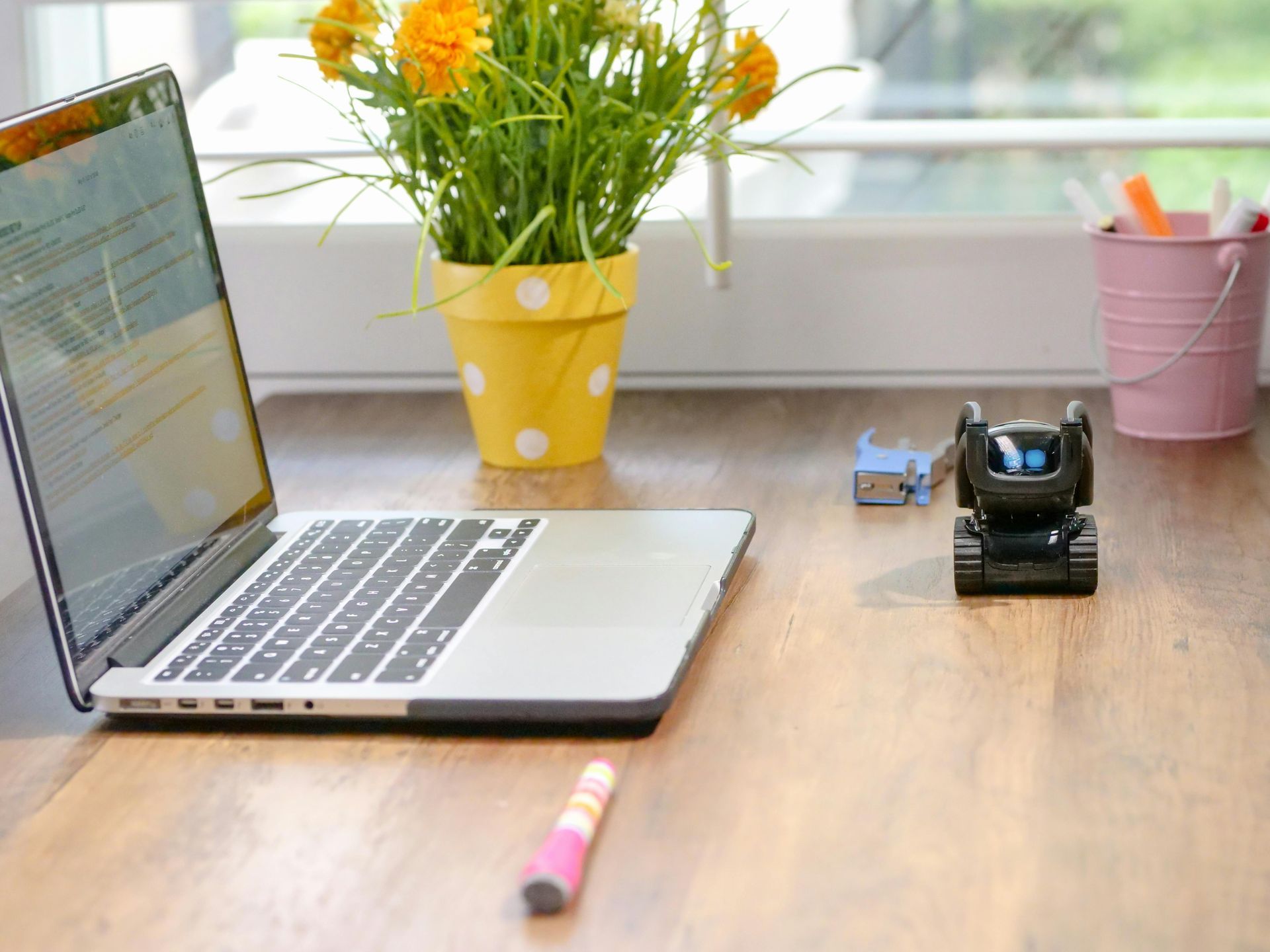 Laptop on a wooden desk with a notebook, flowers in yellow pot, and a tape dispenser by the window
