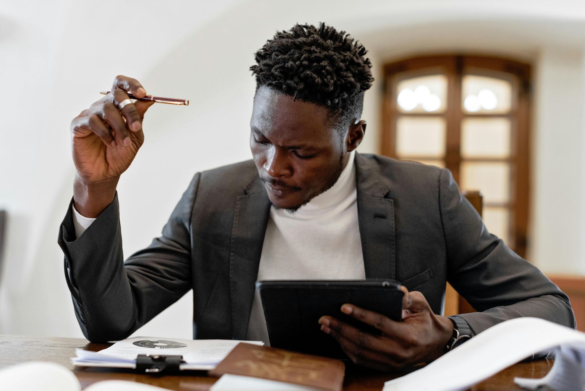 A professional in a grey blazer and white turtleneck reviews information on a tablet while sitting at a desk.