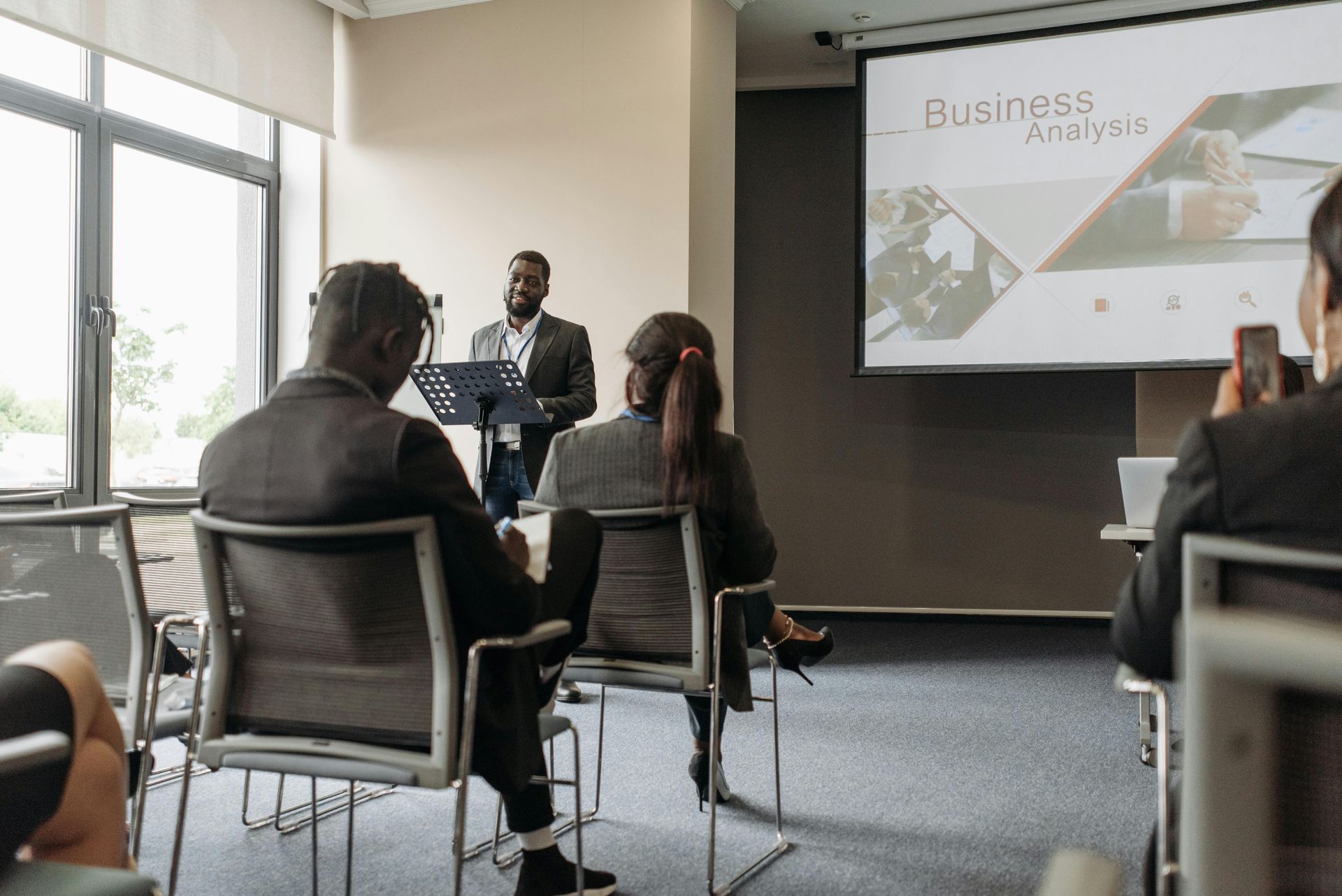 A presenter speaks to an audience in a conference room with a slide titled 
