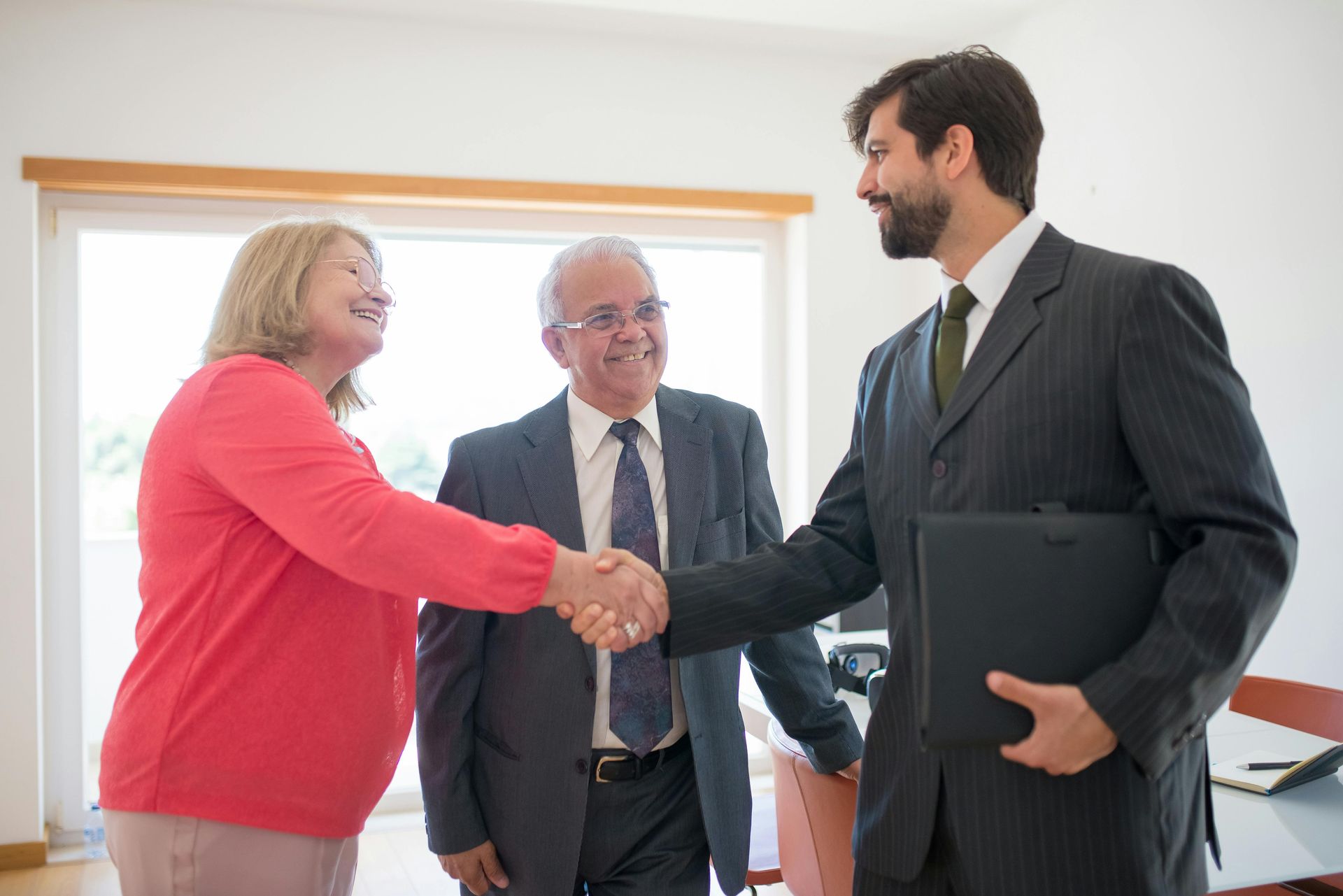 A professional in a suit shakes hands with a person in a pink top, while another person in a suit looks on in an office.