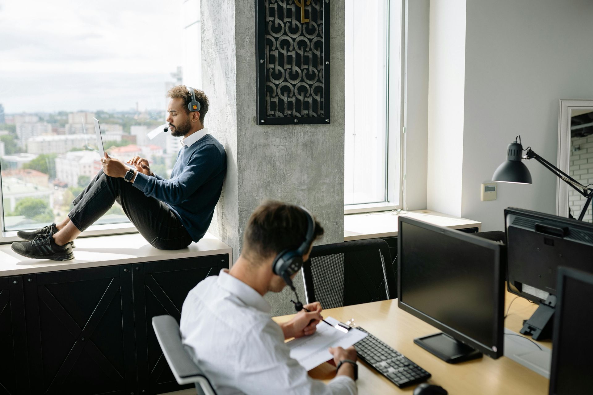 Two people in a modern office, one sitting on a windowsill using a laptop and the other at a desk writing on documents.