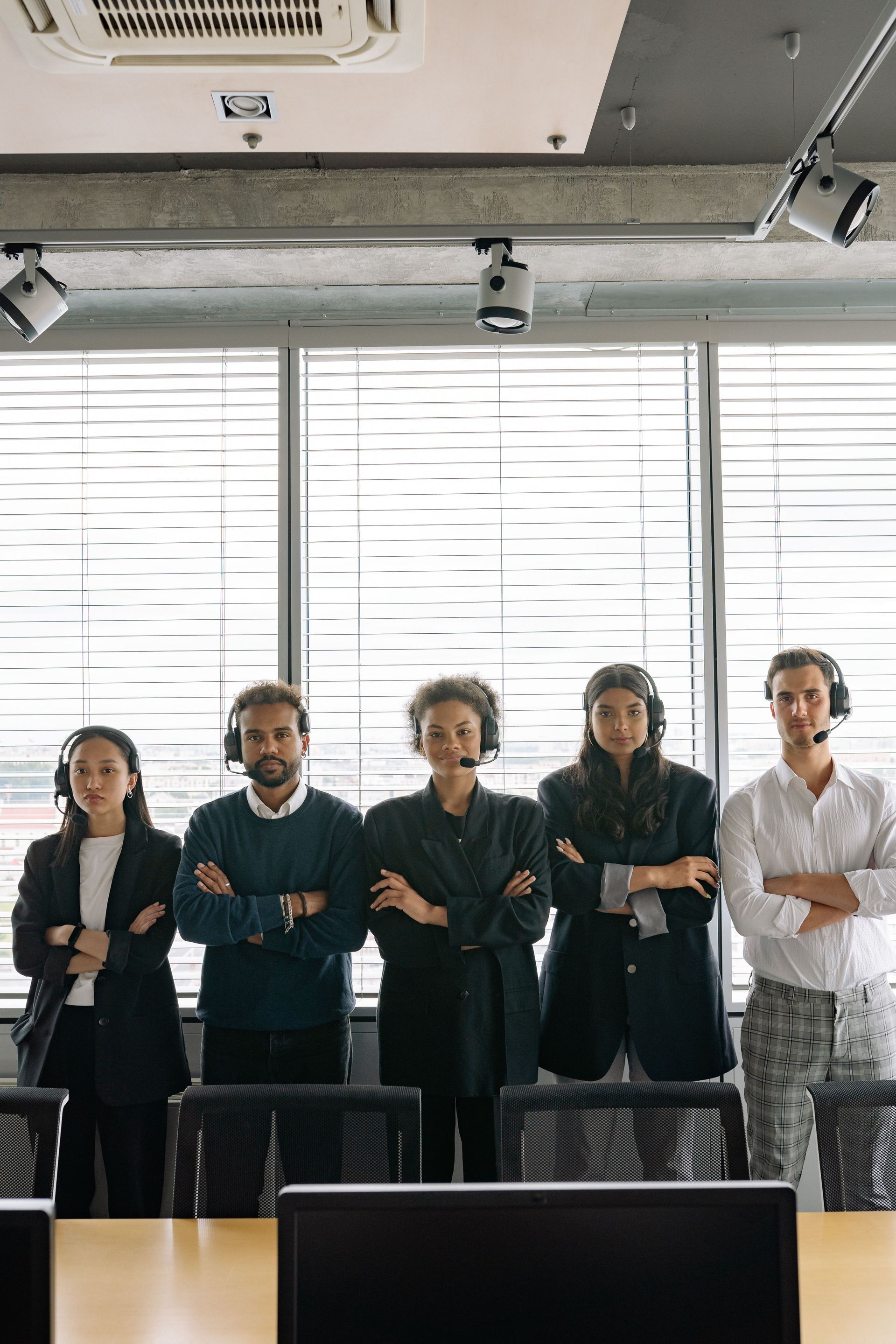 Five people standing in an office with arms crossed, in front of large windows and desks.