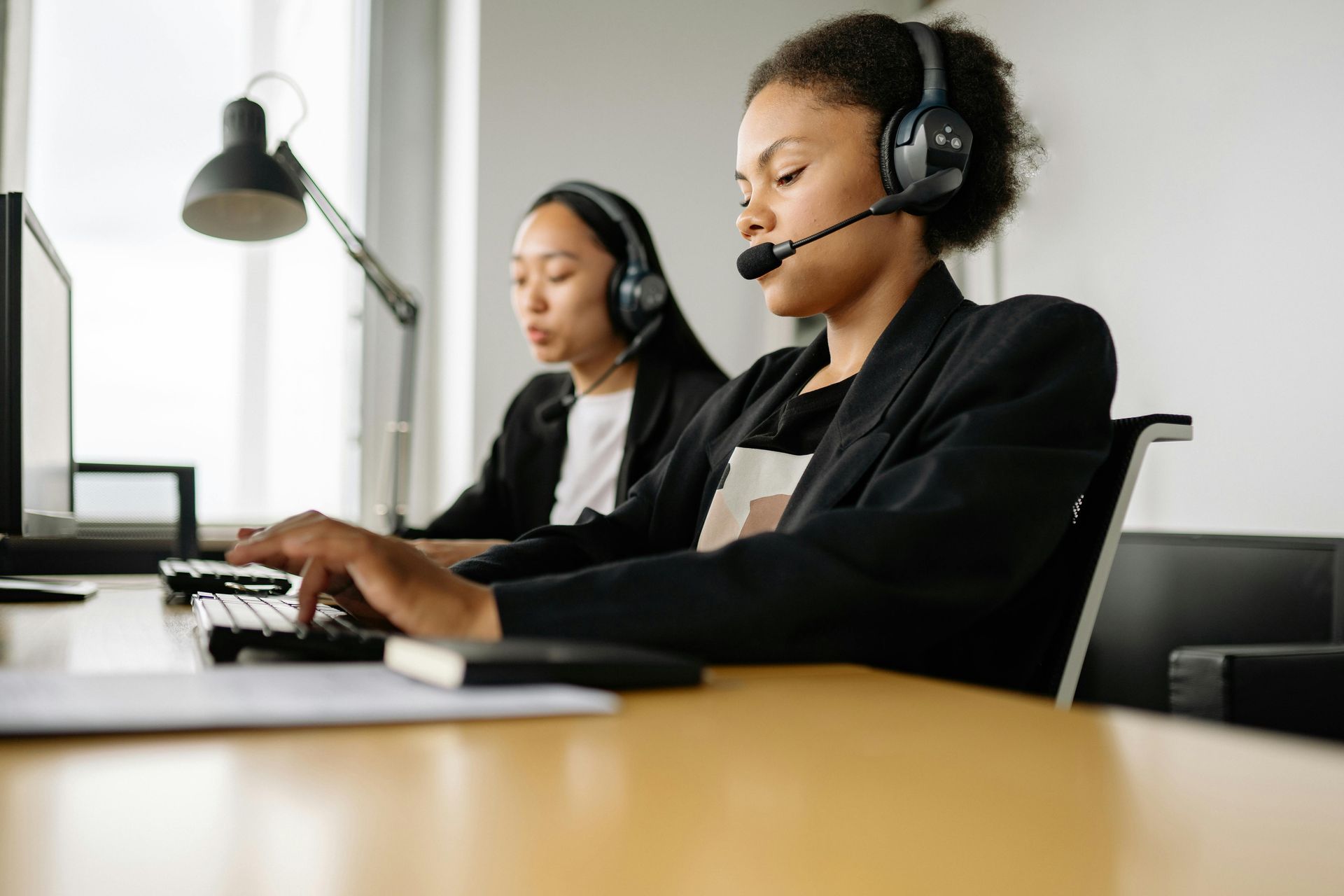 Two people wearing headsets and professional attire work at desks in a bright office, typing on keyboards.
