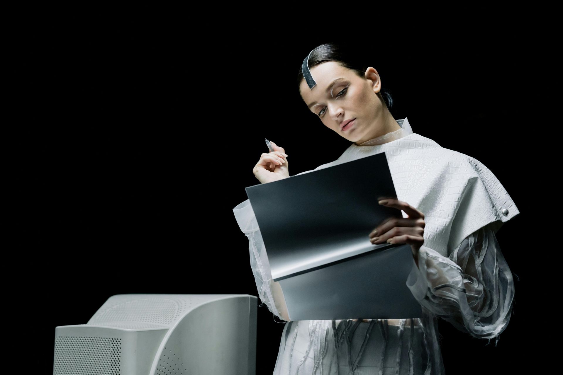 Person in a white outfit holding a folder beside a gray computer, against a black background.
