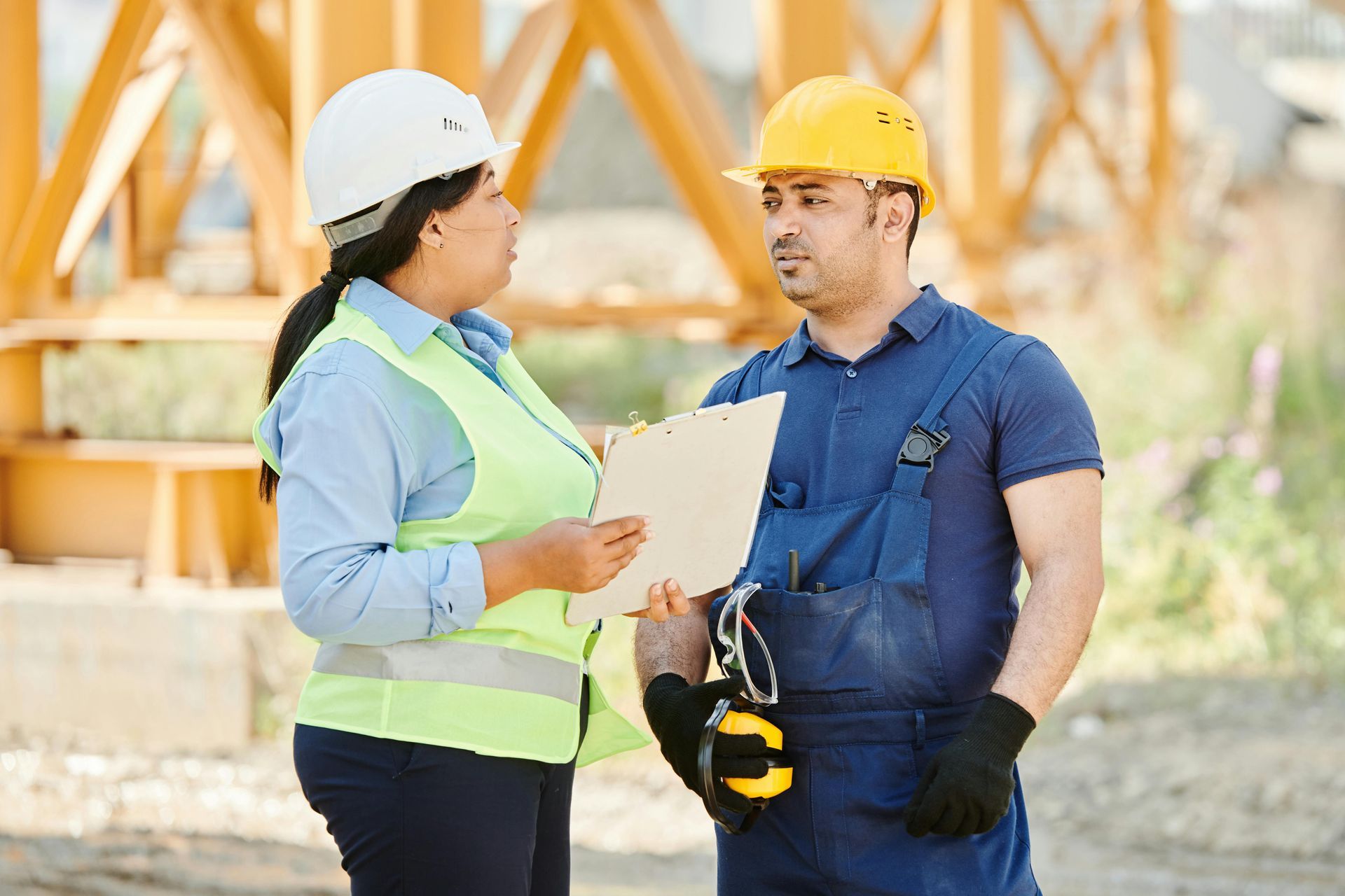 Woman with white helmet and vest discusses documents with a man in blue overalls and yellow helmet at construction site.