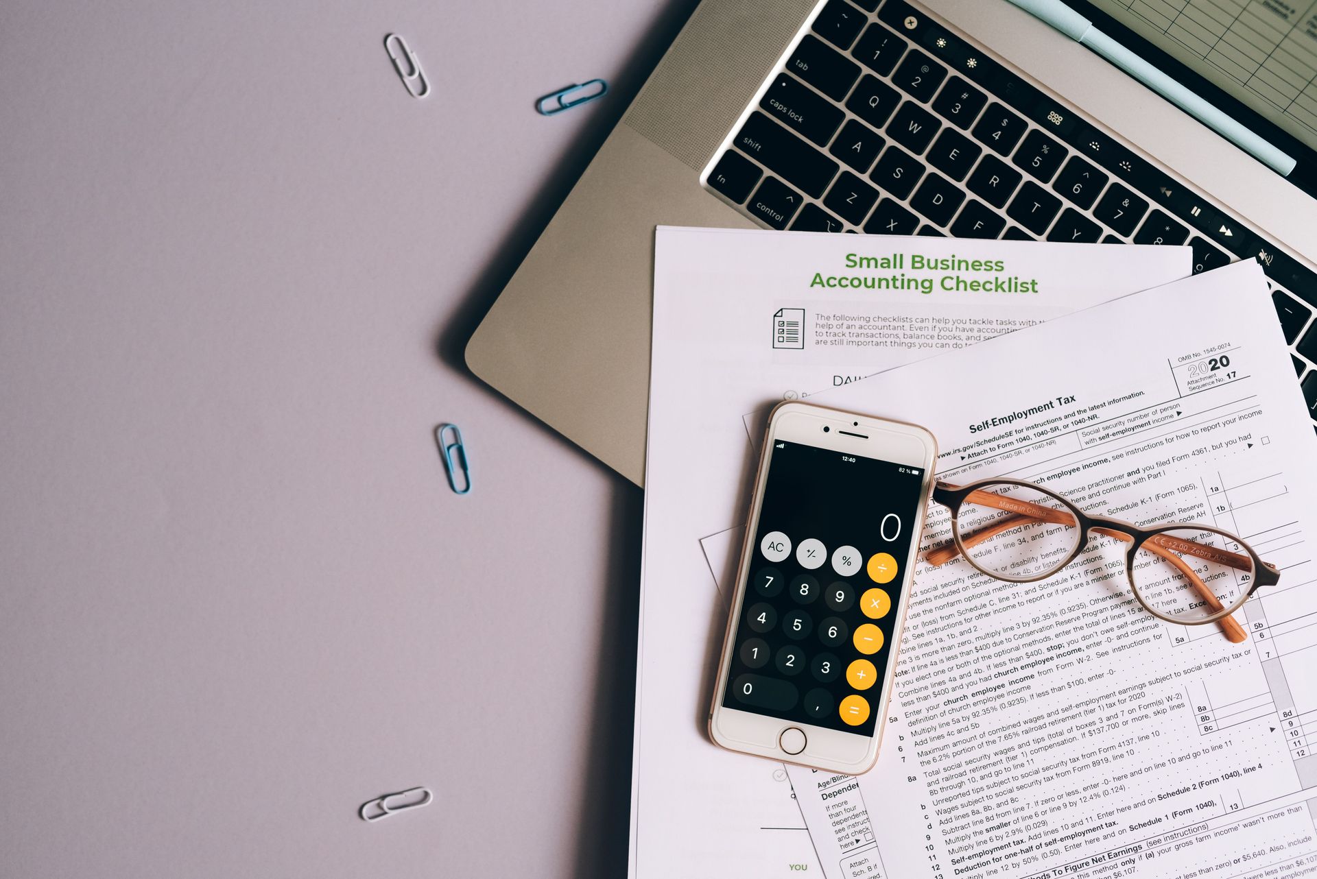 Laptop, calculator, glasses, and paperwork on a desk with paper clips scattered nearby