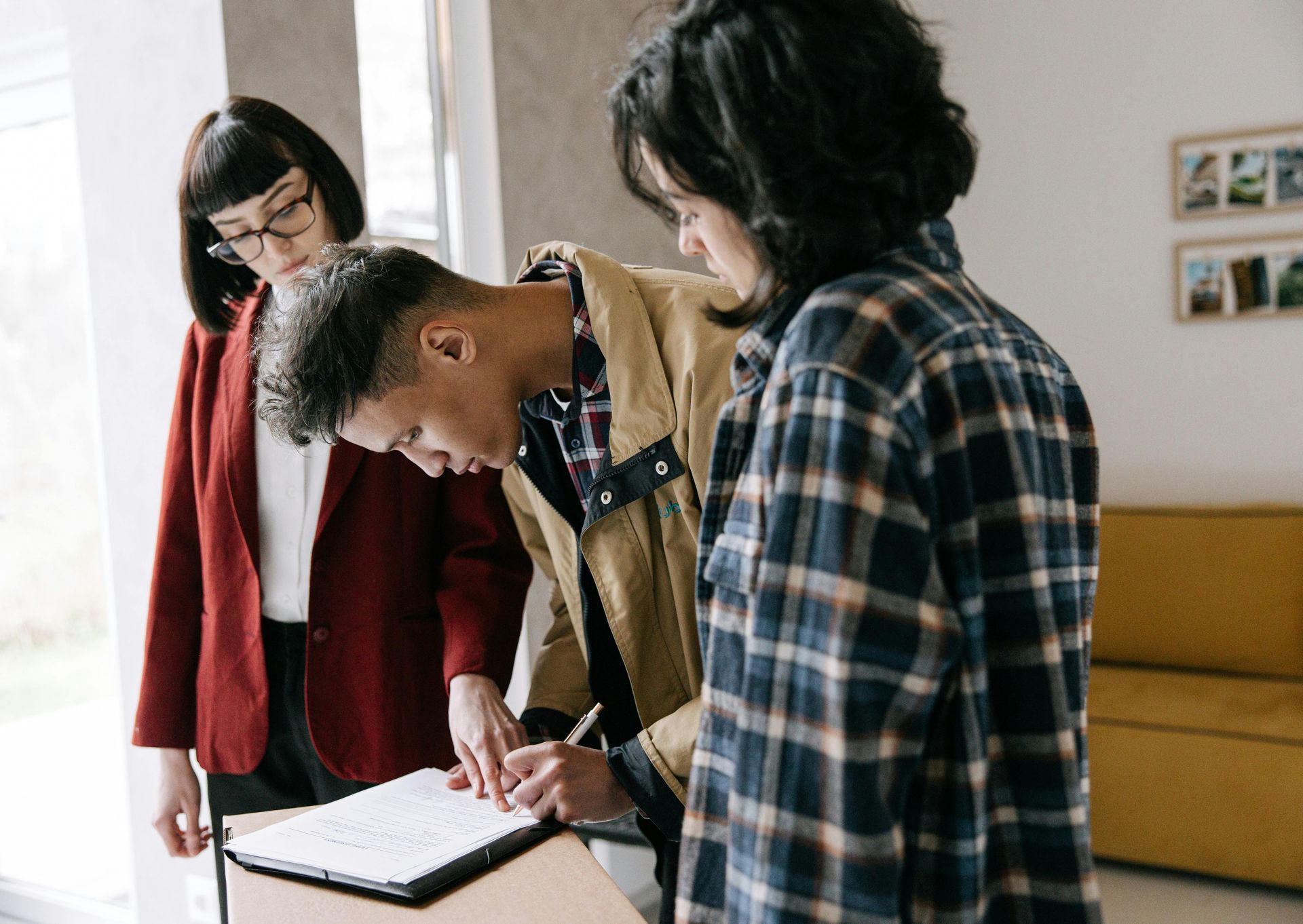 Three people signing a document. A man signs as two others watch. One wears a plaid shirt.