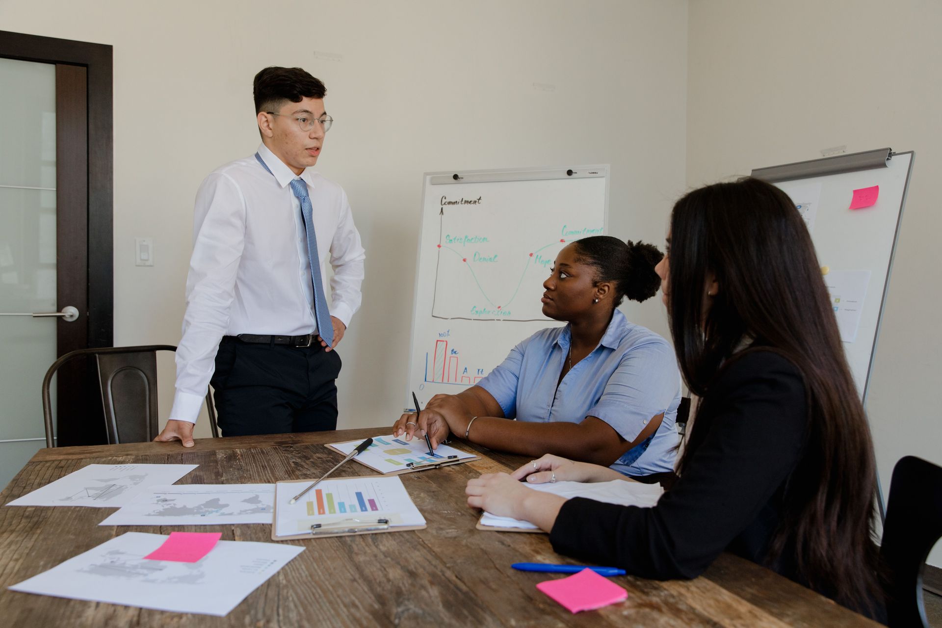 Team meeting in a conference room with papers, charts, and sticky notes on a wooden table.