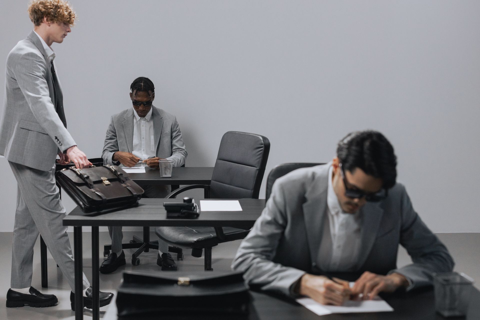 Three people in gray suits work in a minimalist office, one writing at a desk, two seated nearby.