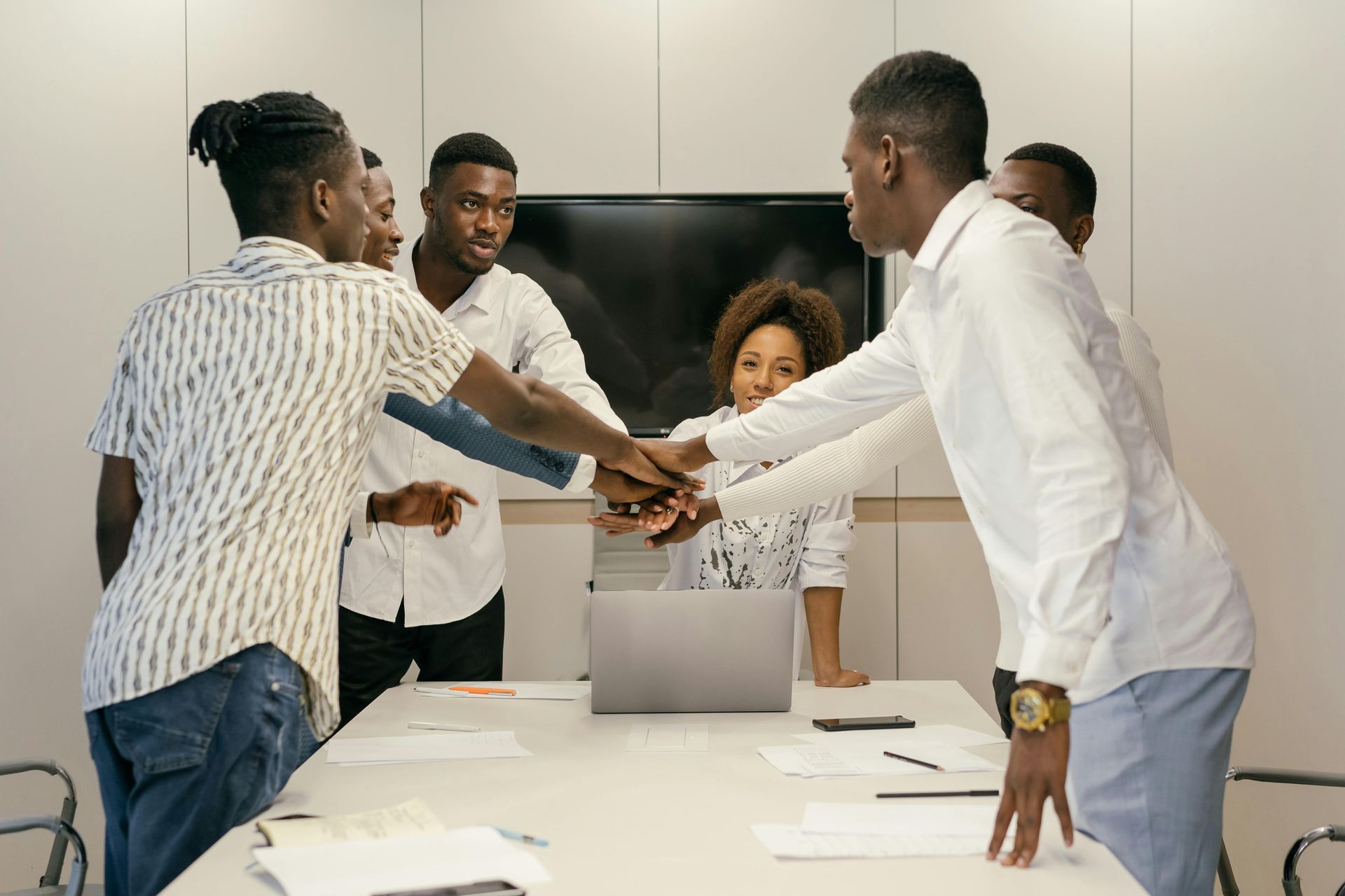 A group of people standing around a conference table in an office, joining their hands together in the center.