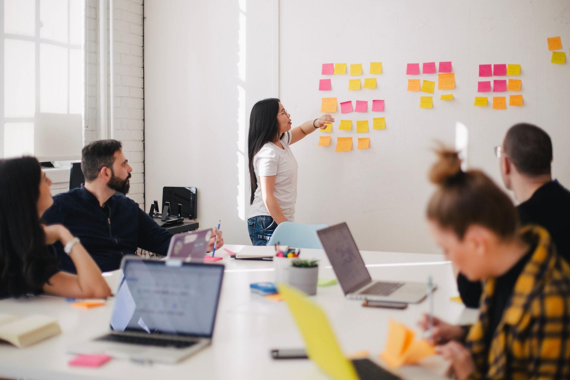 A woman points at sticky notes on a whiteboard, leading a meeting with colleagues around a table with laptops.