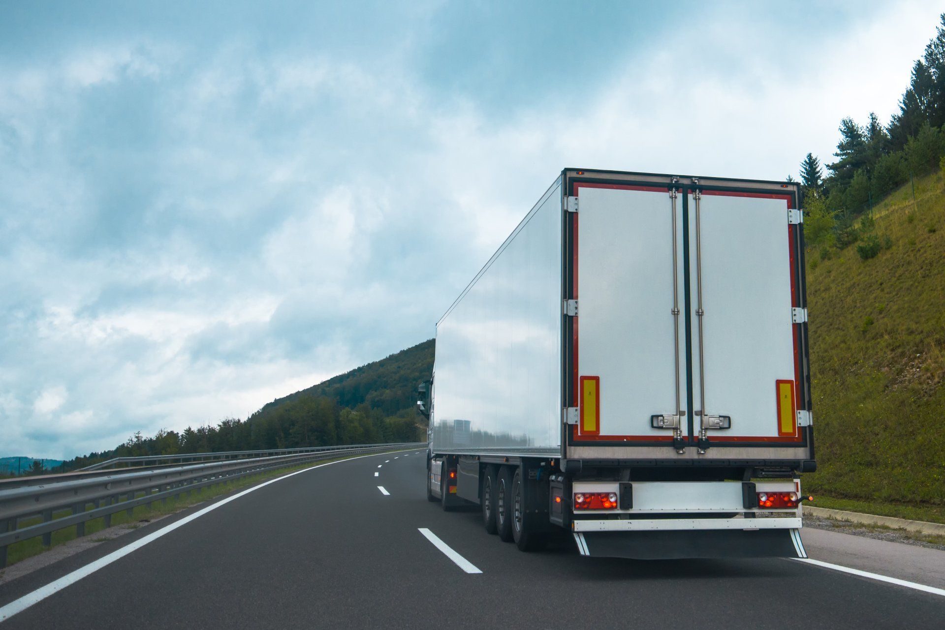 White semi-truck driving on a highway, green hillside and cloudy sky in background.