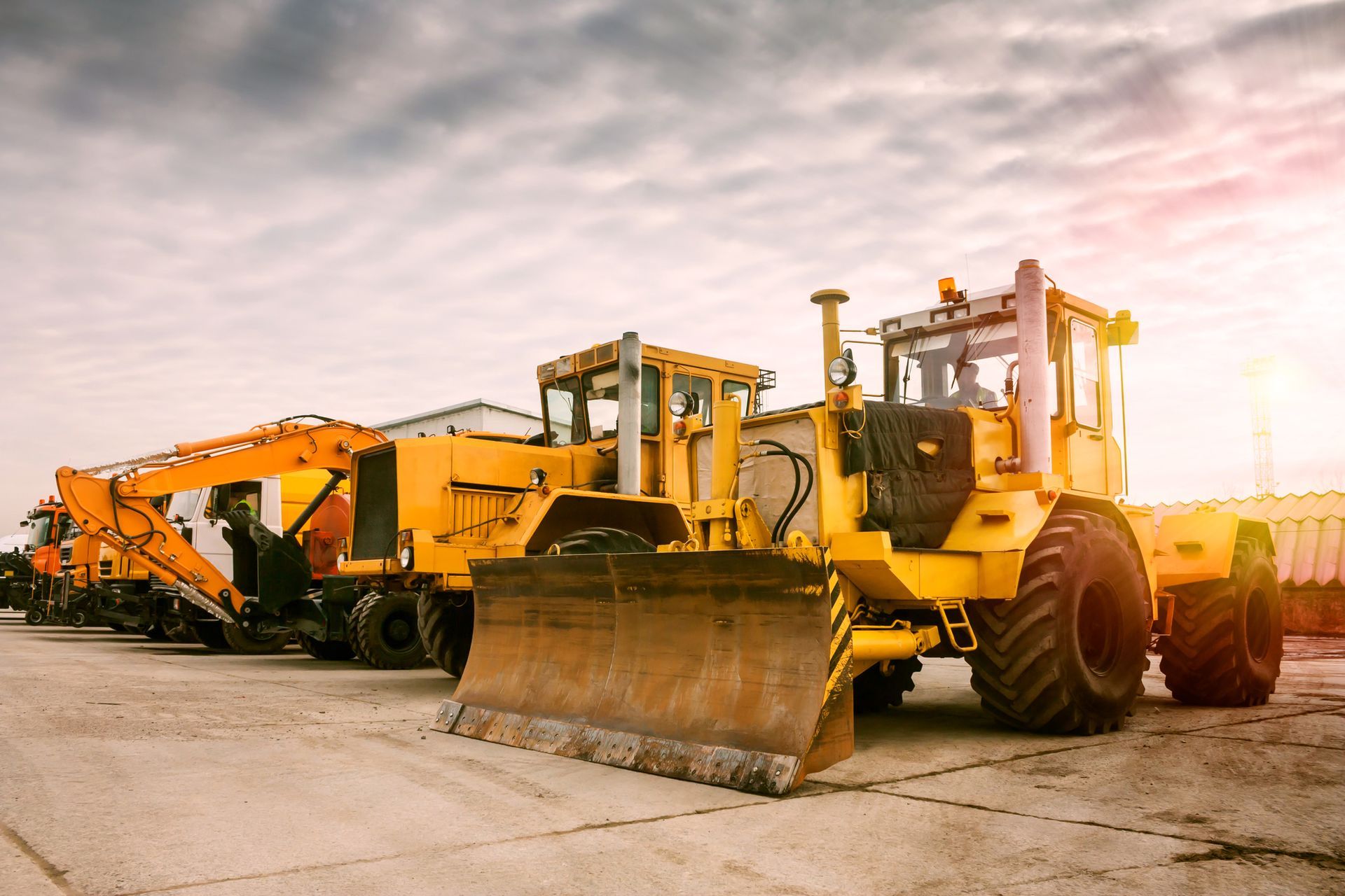Yellow construction vehicles parked on a concrete surface, cloudy sky in the background.