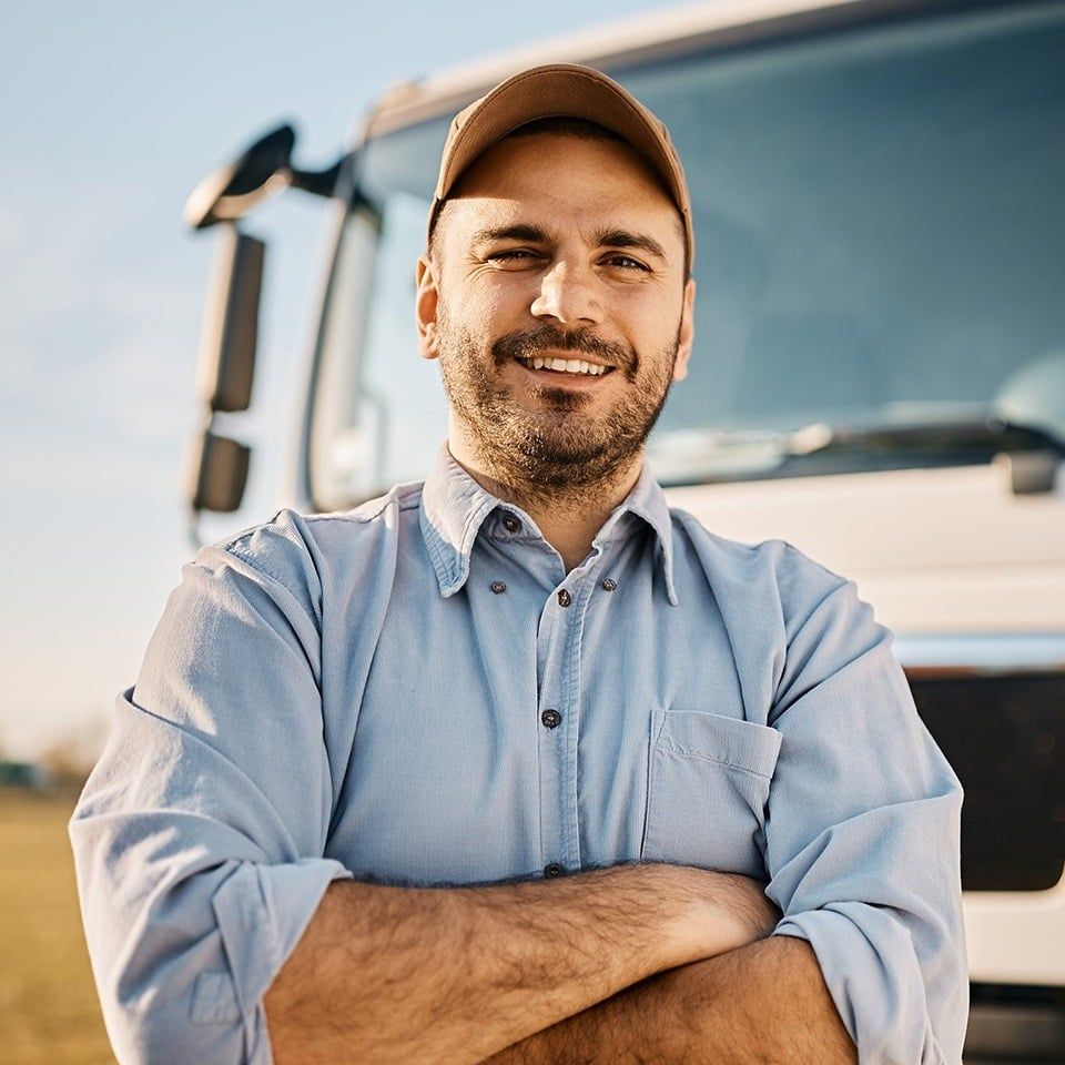 Smiling person with arms crossed, in front of a large truck; wearing a hat and blue shirt.