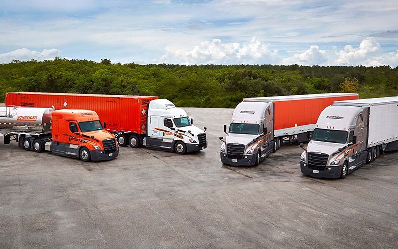 Four semi-trucks parked on concrete; orange and white trailers, forest background.