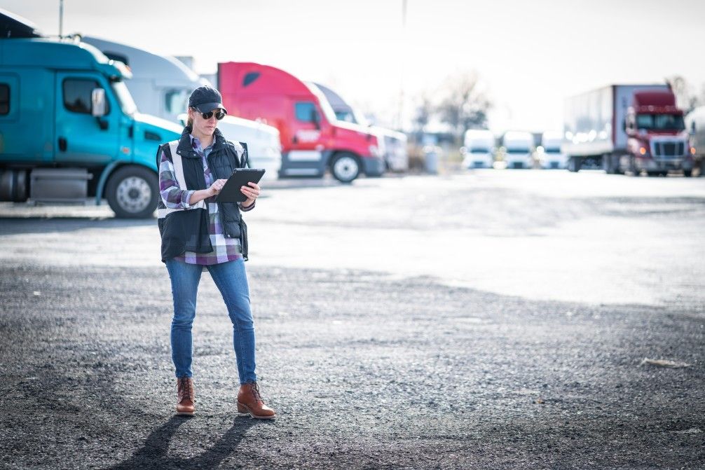 Woman in cap and vest holding tablet, checking trucks in a sunny parking lot.