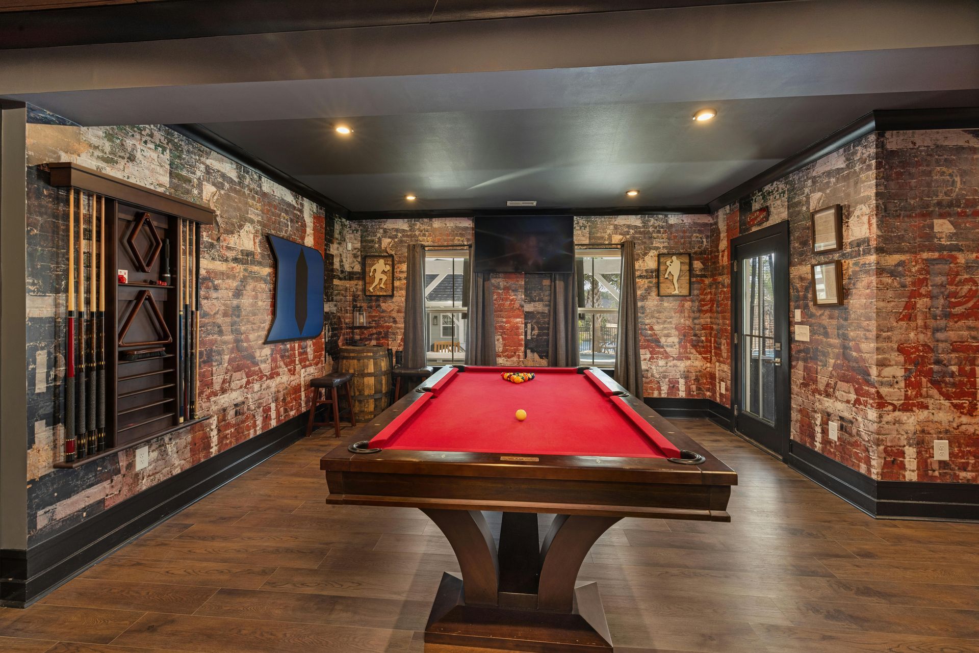 A game room featuring a billiards table with a red felt top, wood-paneled walls with vintage print, and cue rack storage.