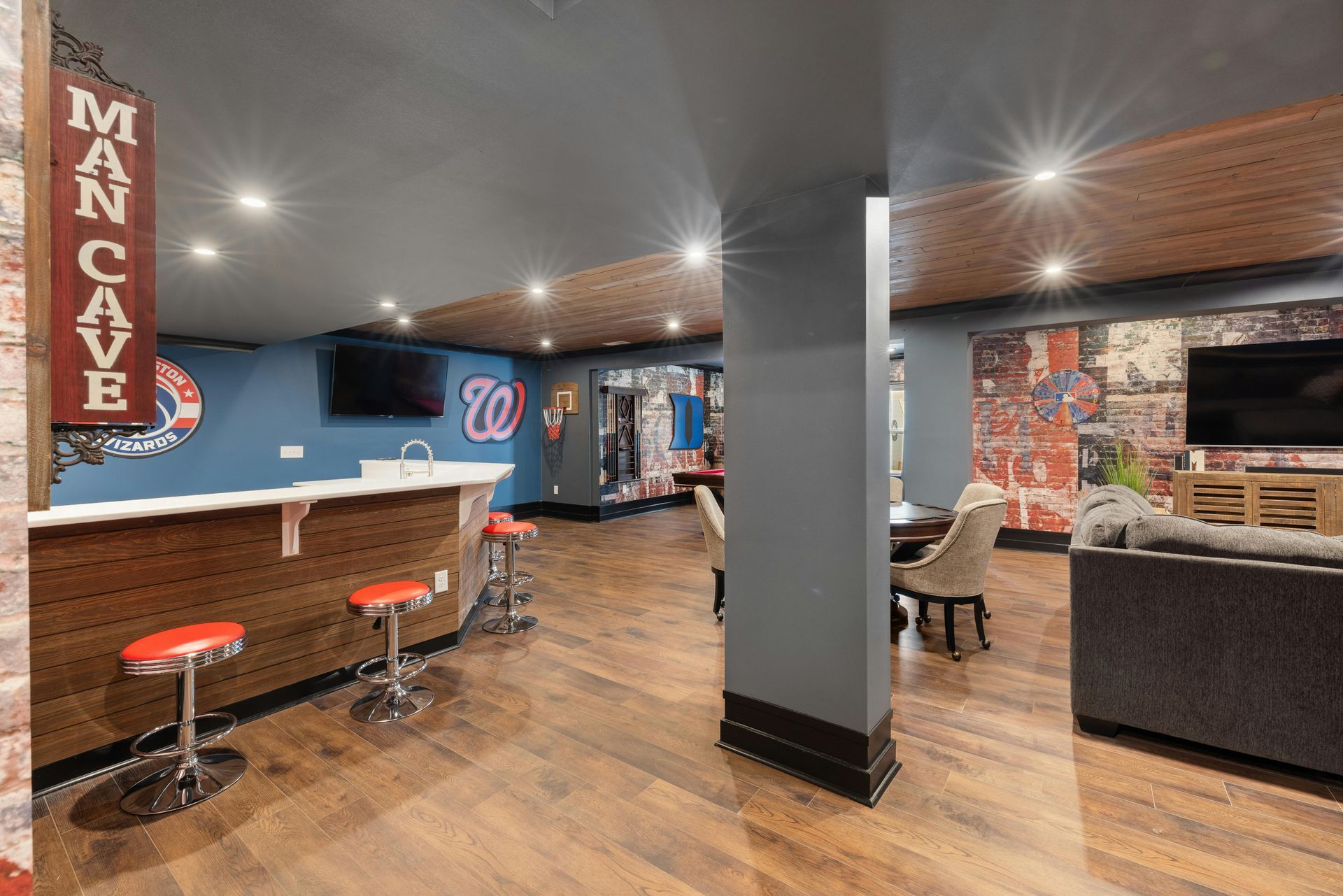 A basement man cave with a bar, red stools, a dark support pillar, a seating area with a sofa, and sports decor.