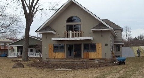 Two-story tan house with boarded windows and a small balcony in a bare yard