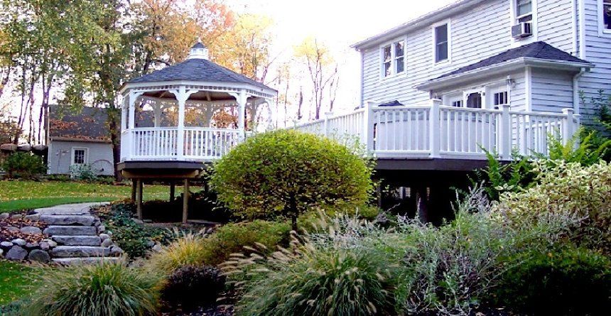 Backyard with white gazebo on a deck beside a house, surrounded by lush green shrubs and landscaping.