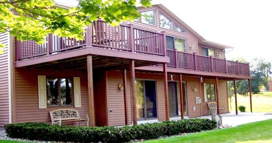 Two-story house with red siding, large wooden deck, and manicured lawn surrounded by trees
