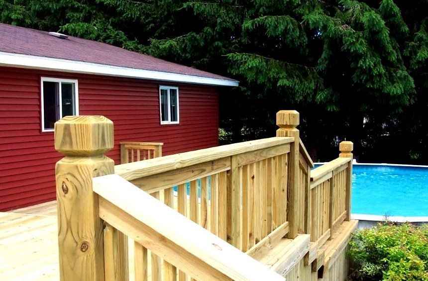 Wooden deck railing beside a red house, with a blue swimming pool and green trees in the background.