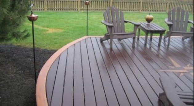 Curved wooden deck with Adirondack chairs beside a green lawn and fence after rain