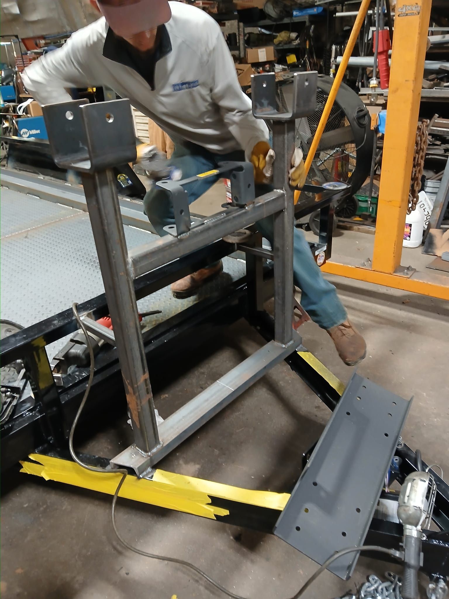 Person installing metal frame on a trailer, wearing work clothes. Shop setting, metal equipment.