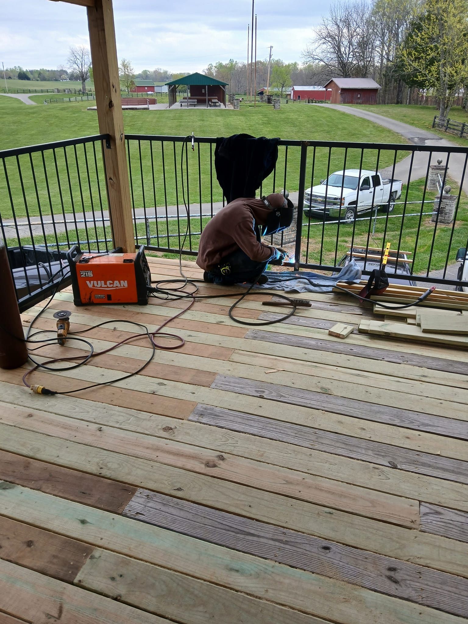 Person welds metal railing on wooden deck, welding machine nearby. Rural setting with buildings and vehicles.