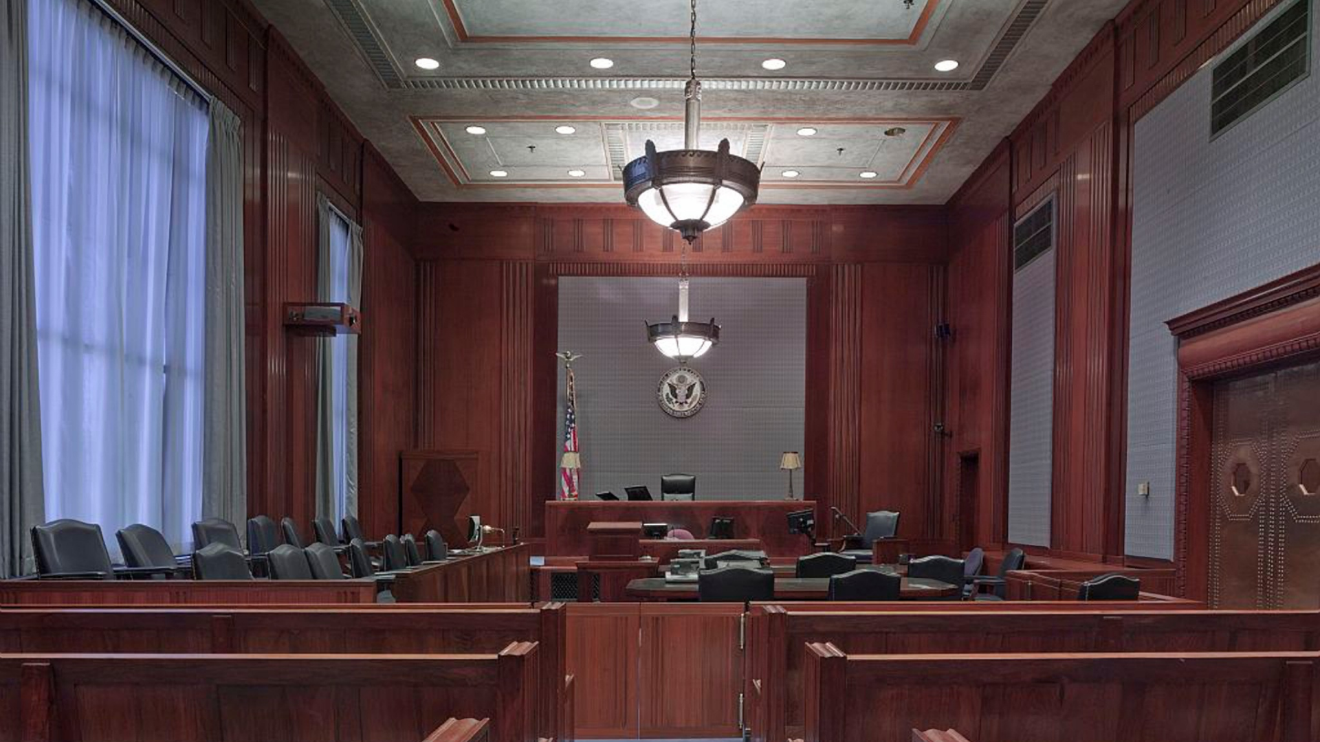 Empty courtroom with wooden benches, red walls, and a judge’s bench beneath a clock and chandelier.
