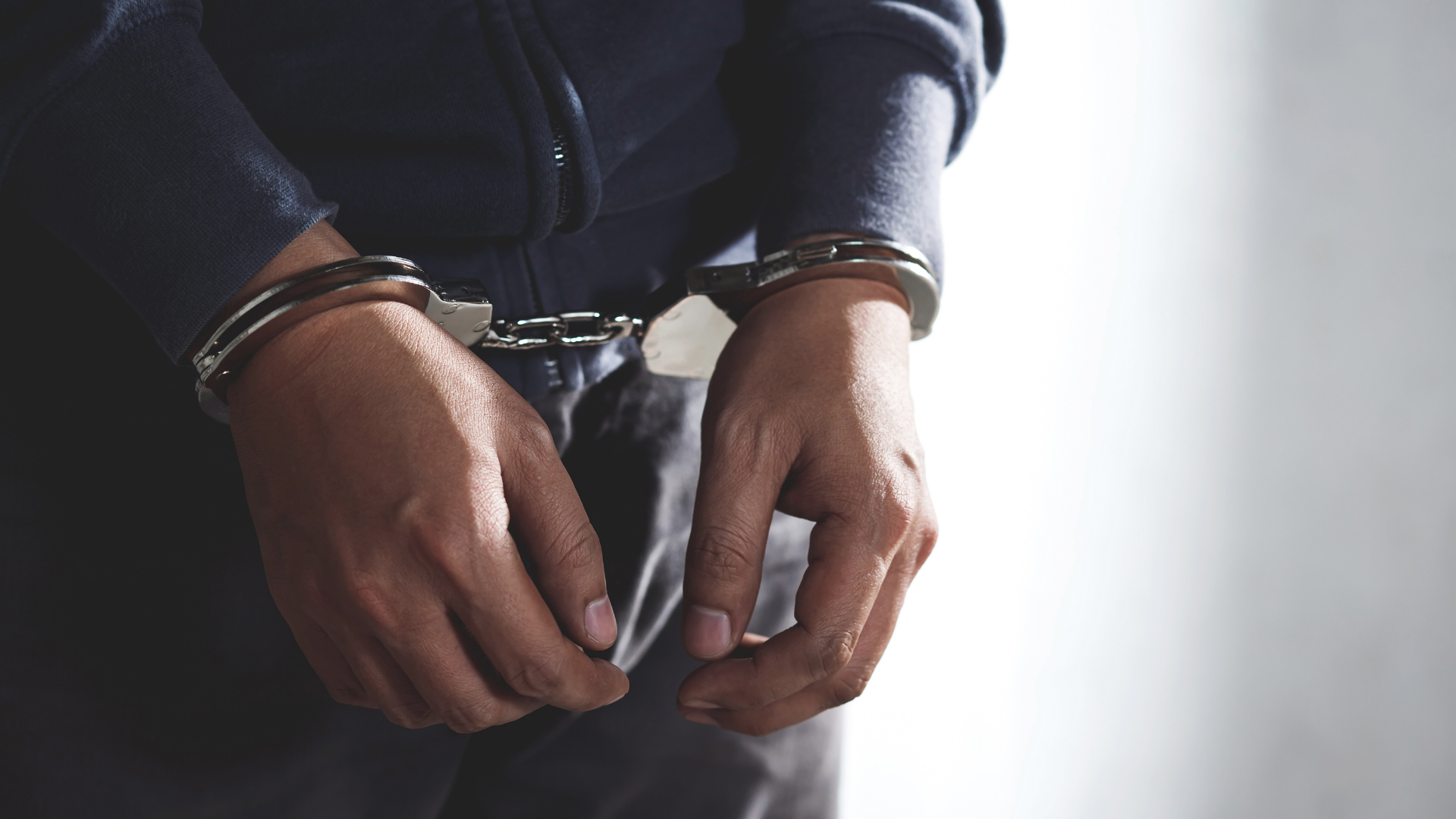 Person in handcuffs, hands restrained at waist in a dark jacket.