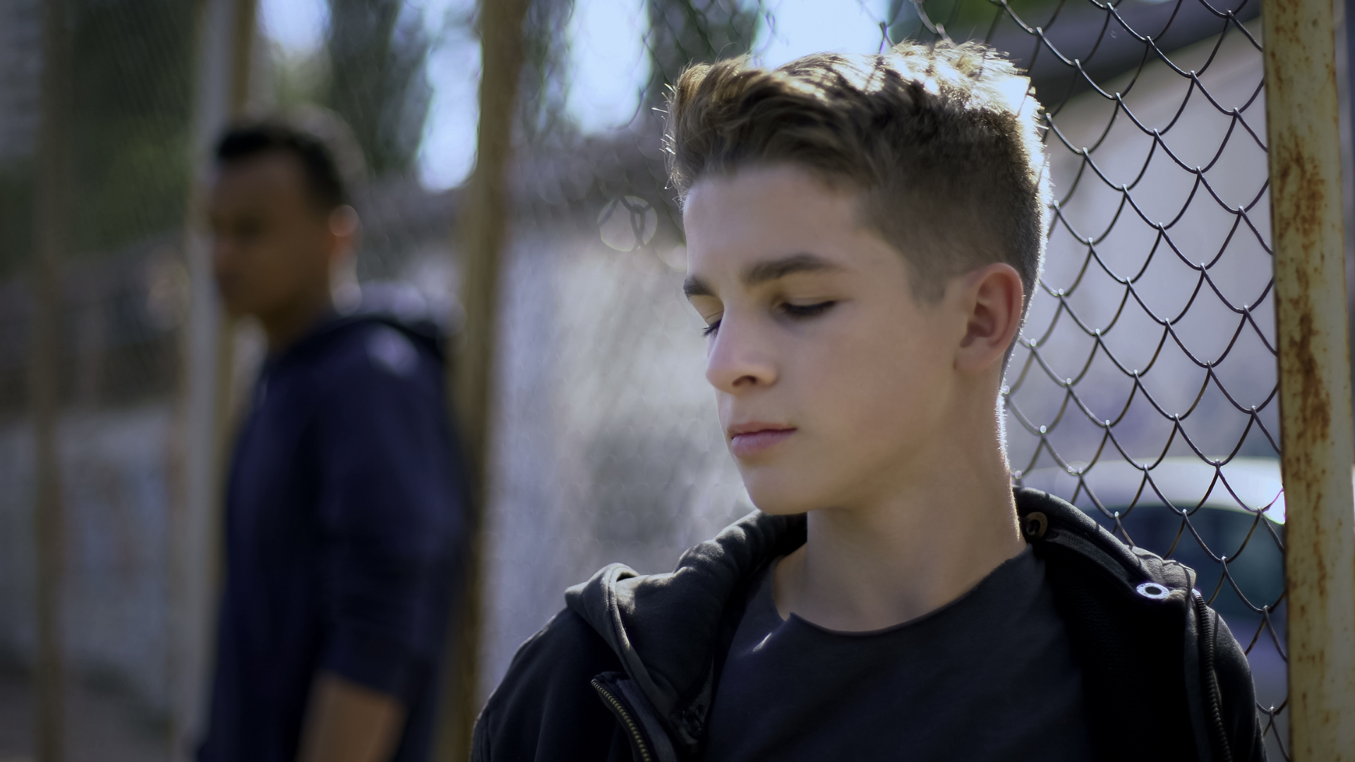 Teen standing by a chain-link fence with eyes closed, another person blurred in the background