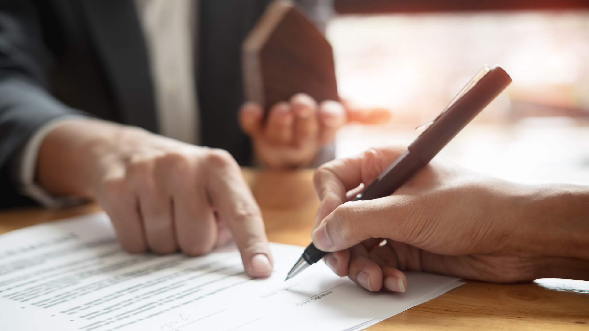 Two people signing a document, one pointing at the paper with a pen in a bright office.