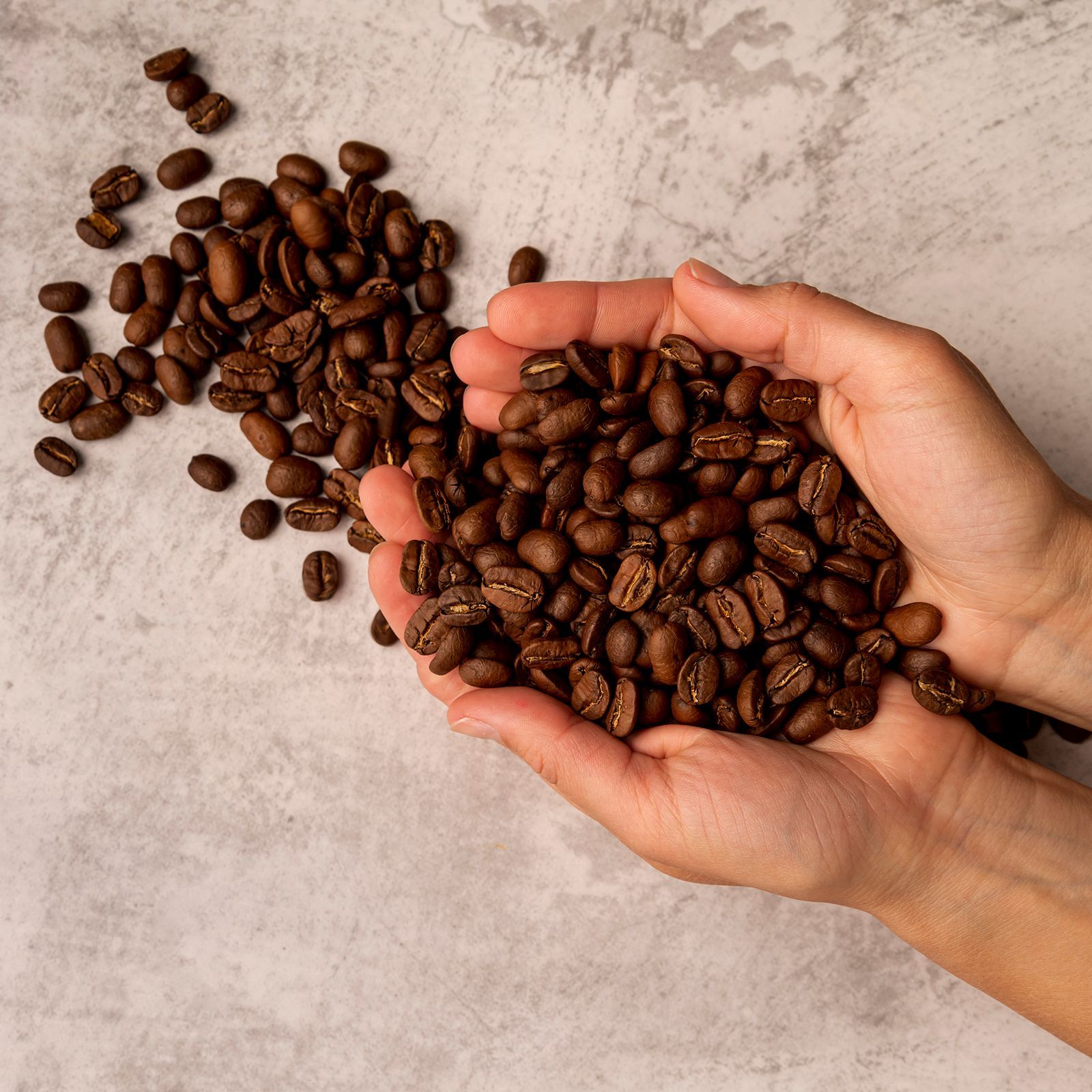 Manos sosteniendo una pila de granos de café tostados, con más granos esparcidos sobre una superficie gris.
