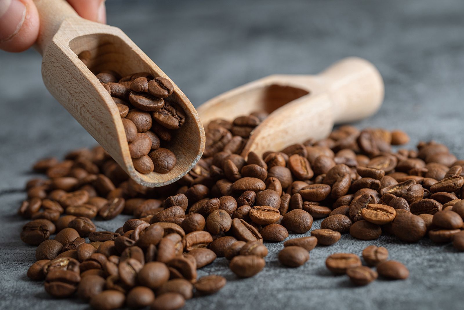 Granos de café derramándose desde dos cucharas de madera sobre una superficie oscura.