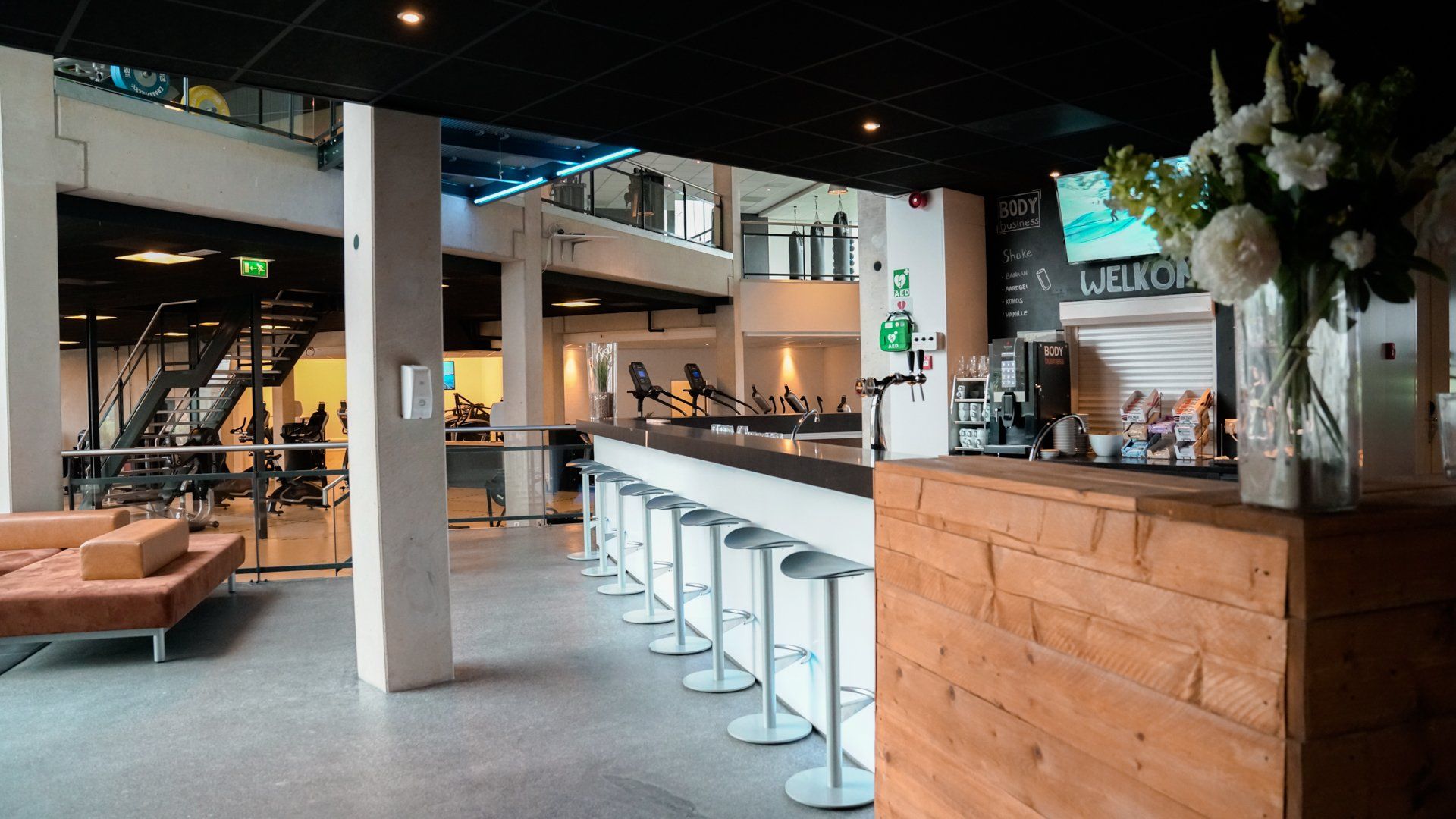 Barruimte in een sportschool: witte barkrukken, houten toonbank, koffiemachine, bloemen en mensen op