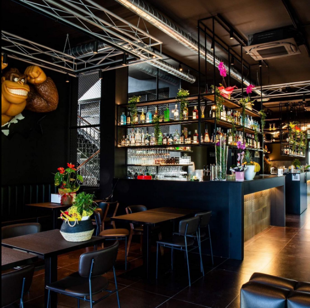 Interni di un bar moderno con scaffali pieni di bottiglie, tavoli scuri e luci soffuse e calde.