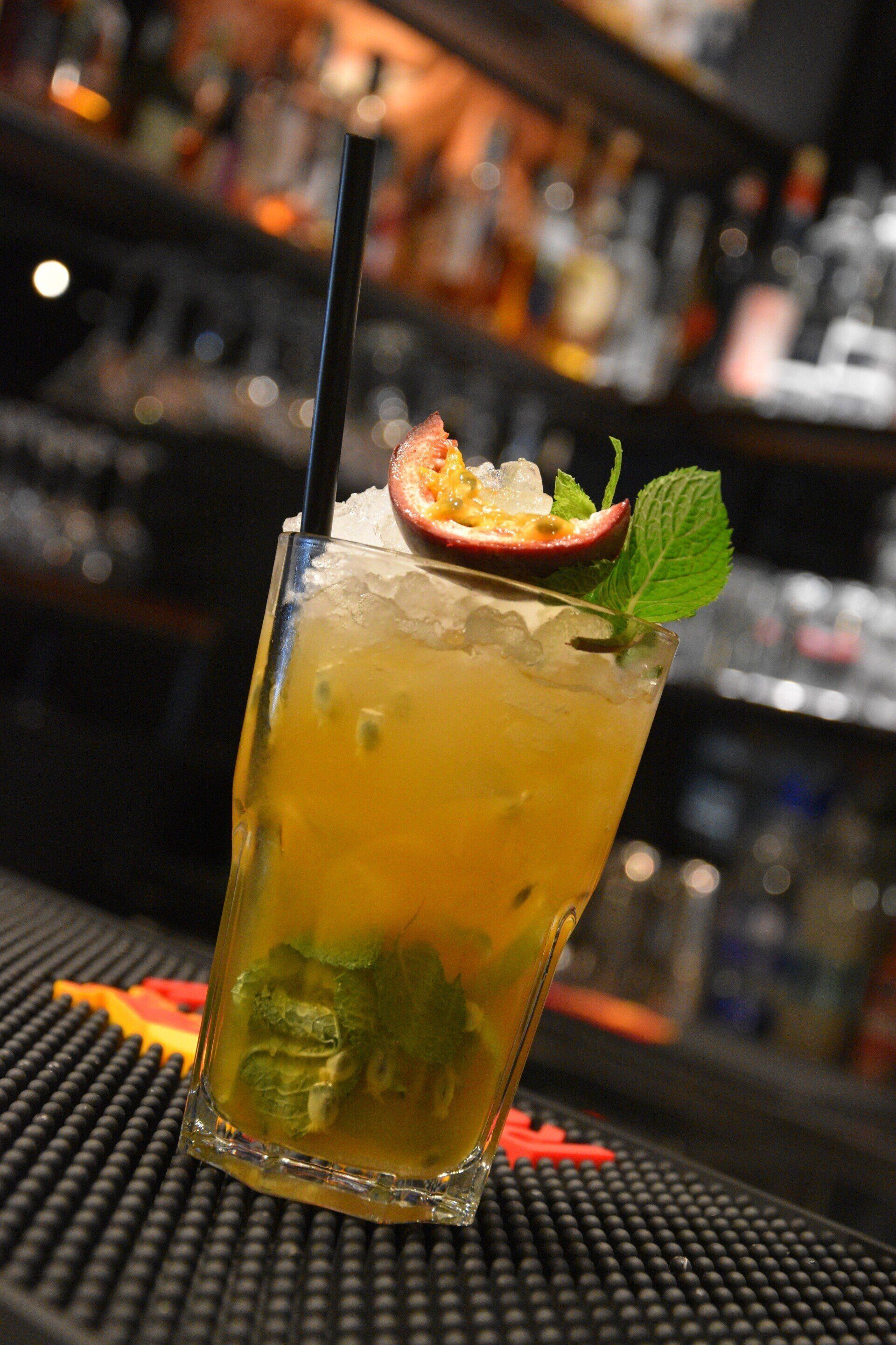 Un cocktail alto e giallo, guarnito con menta e frutto della passione, servito su un bancone da bar.