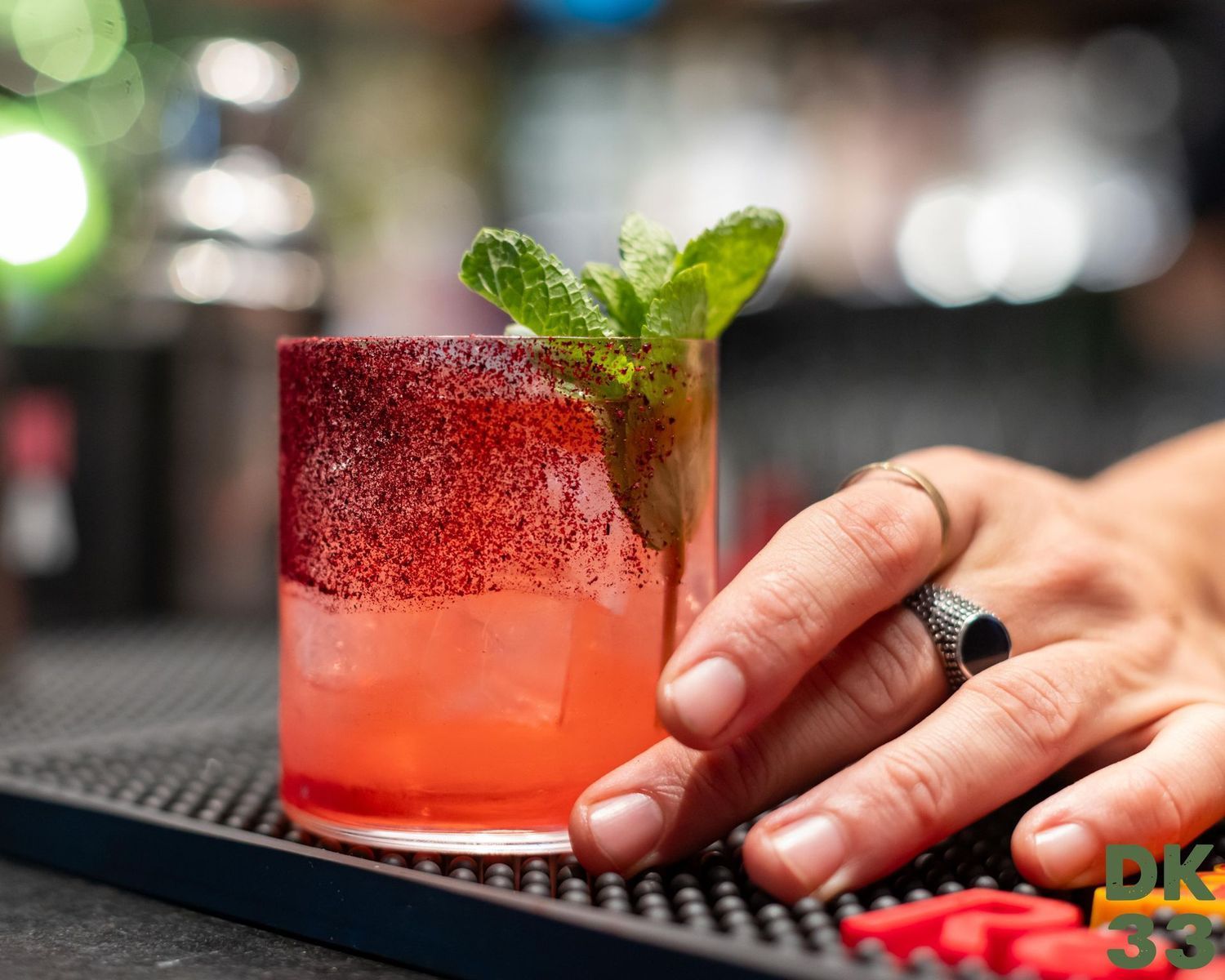 Una mano posiziona un cocktail rosso con menta sul bancone del bar.